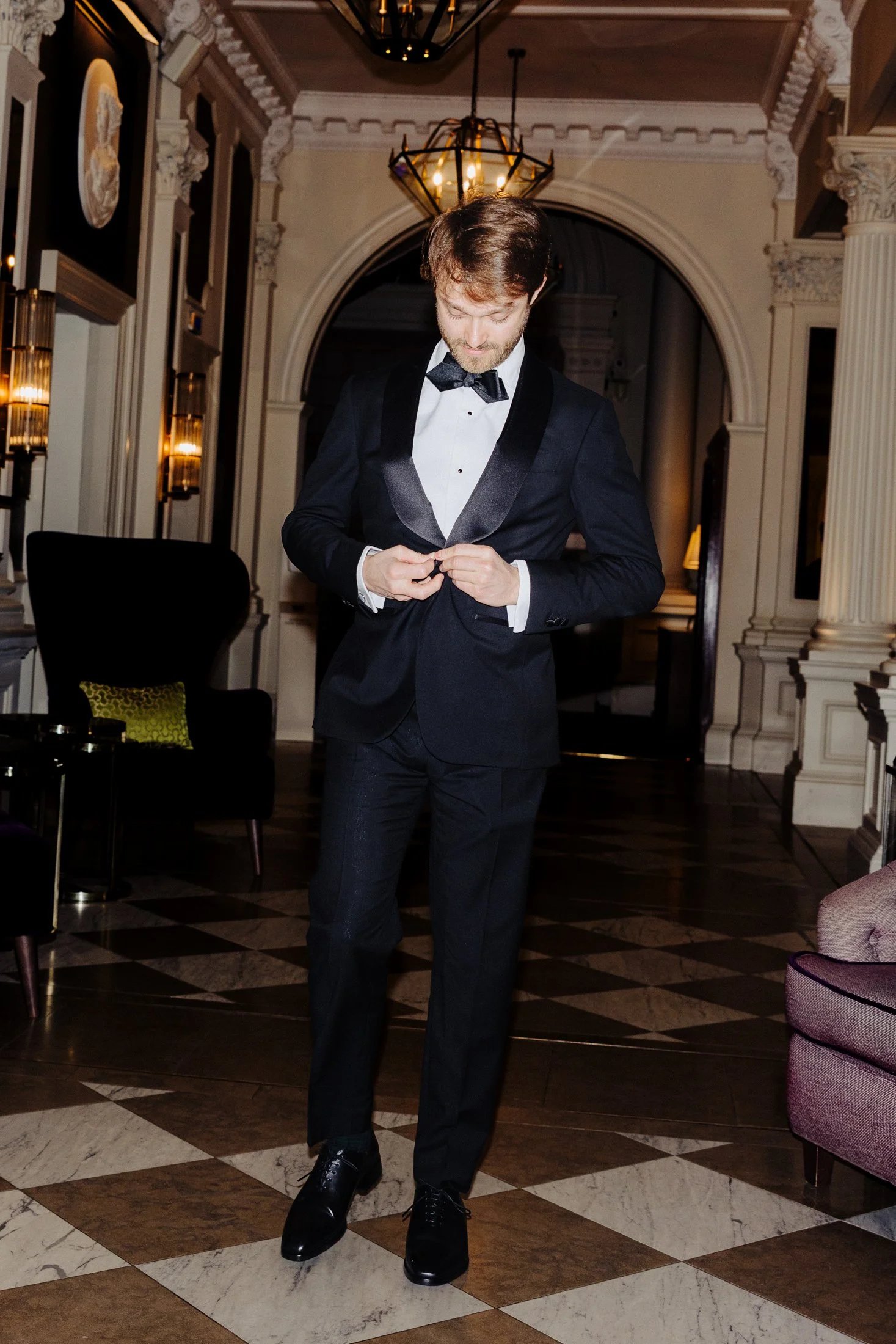 A man in a tuxedo adjusting his jacket in an elegant, ornately decorated room with high ceilings, chandeliers, and marble floors.