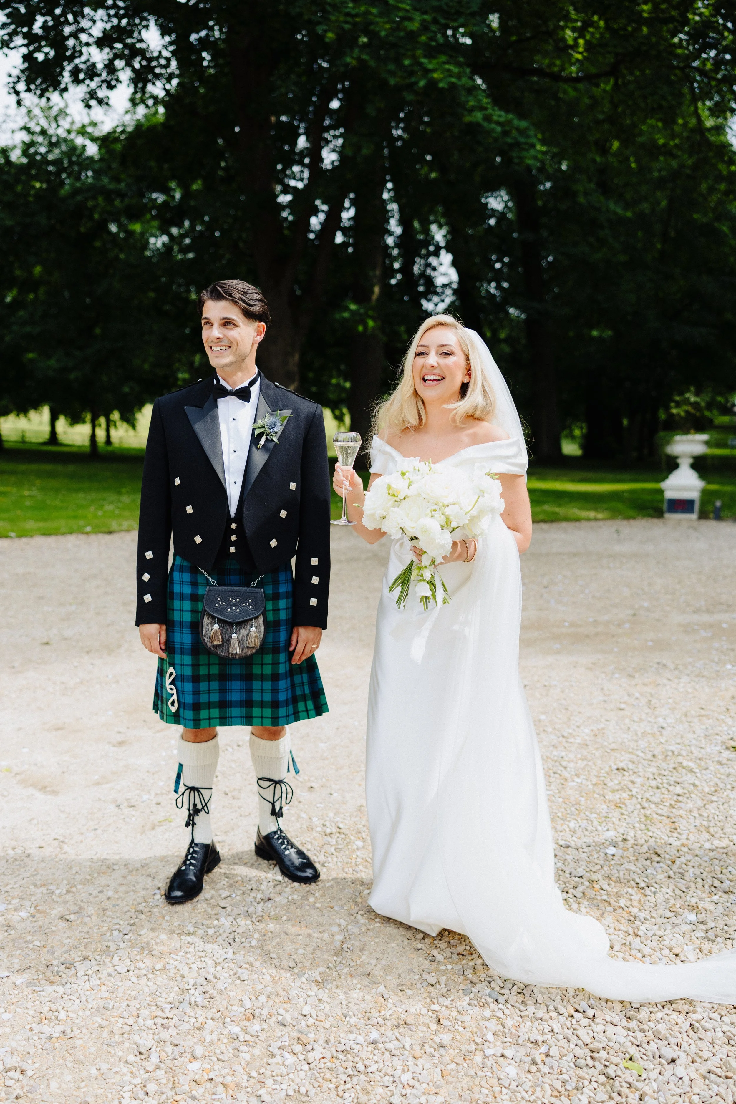 A newlywed couple stands outdoors on a gravel path with greenery and trees in the background, smiling and looking happy. The groom wears a traditional Scottish kilt and jacket, while the bride wears a white wedding dress and holds a bouquet of white 