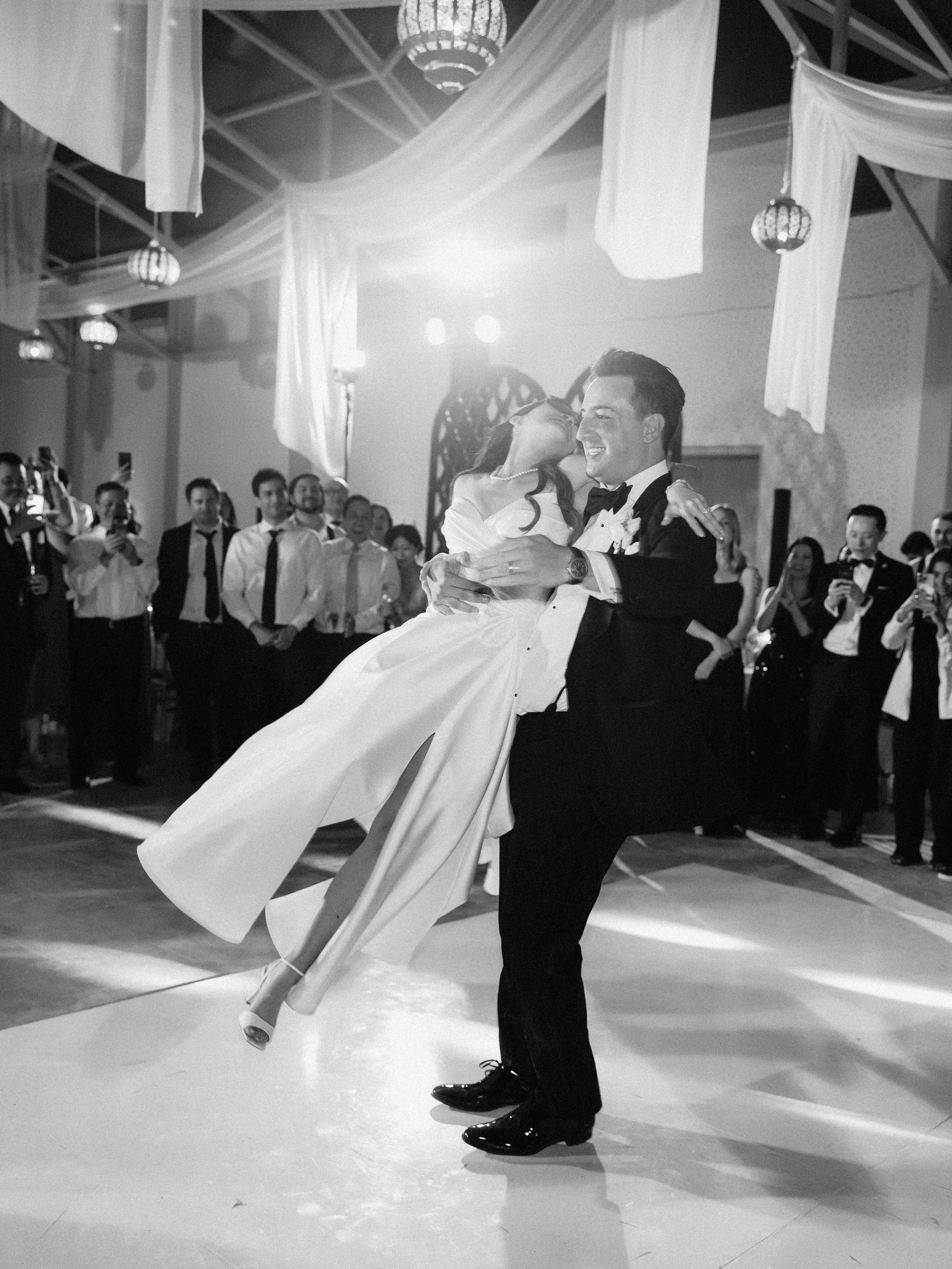 A groom in a tuxedo is spinning a bride in a wedding gown during their first dance at a wedding reception, with guests watching and taking photos.