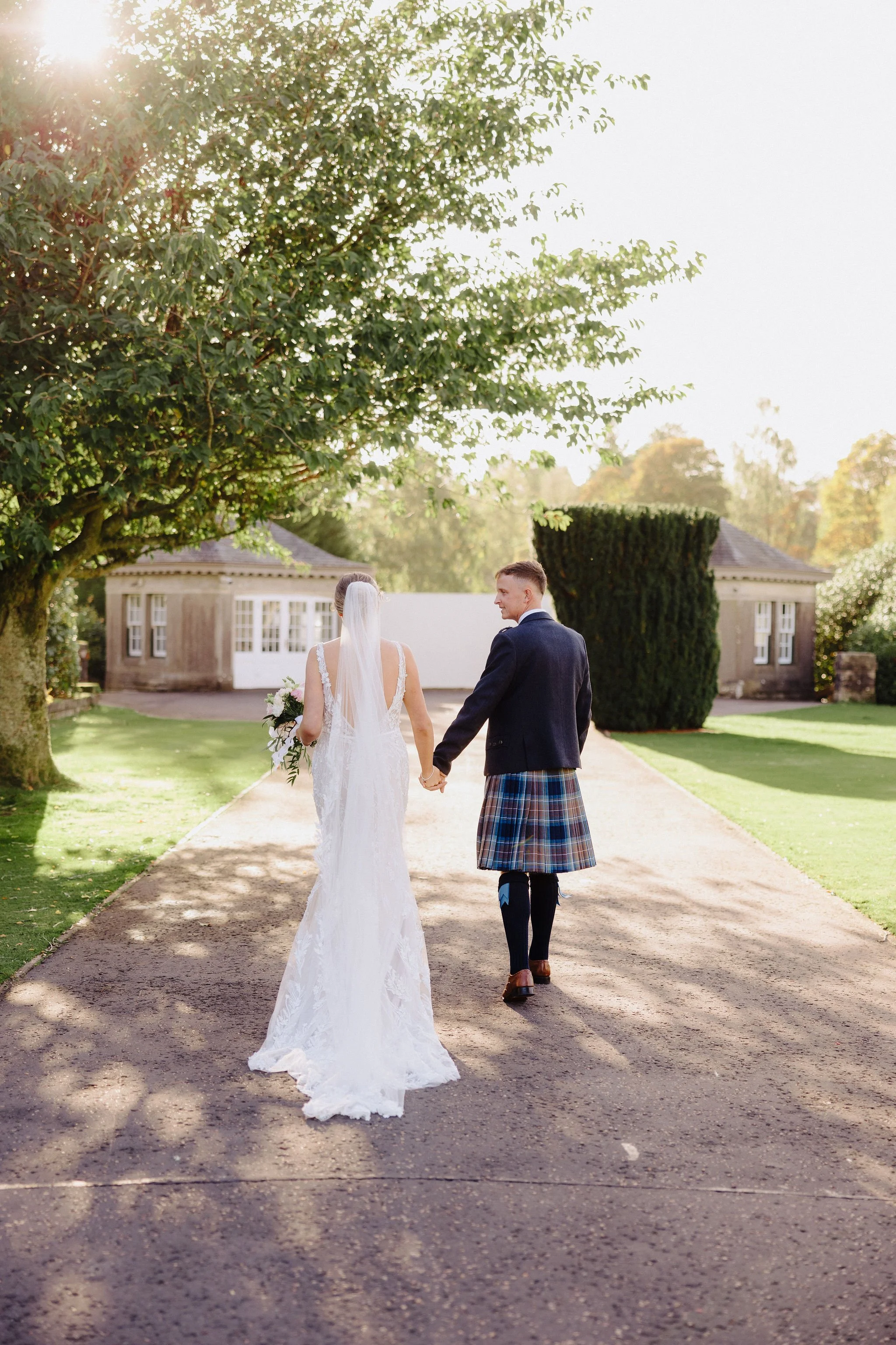 Bride and groom walking hand-in-hand on a park pathway during a wedding, with trees and small buildings in the background, sunlight filtering through the leaves.