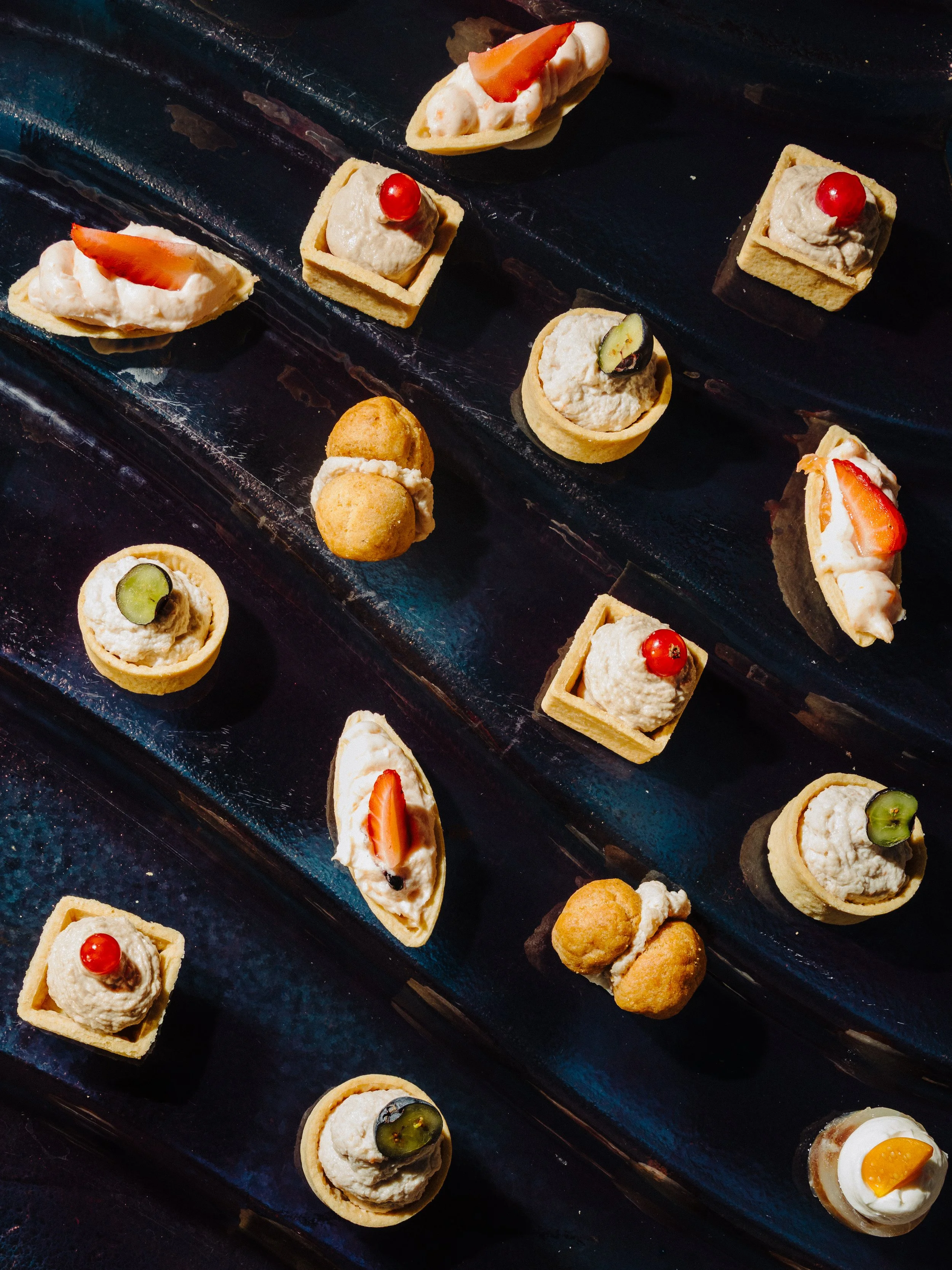 Assorted miniature appetizers on a black serving platter, including tartlets with cream and strawberries, square pastry bites with cucumber and cream, and small round puff pastries filled with cream and topped with a cherry or a strawberry.