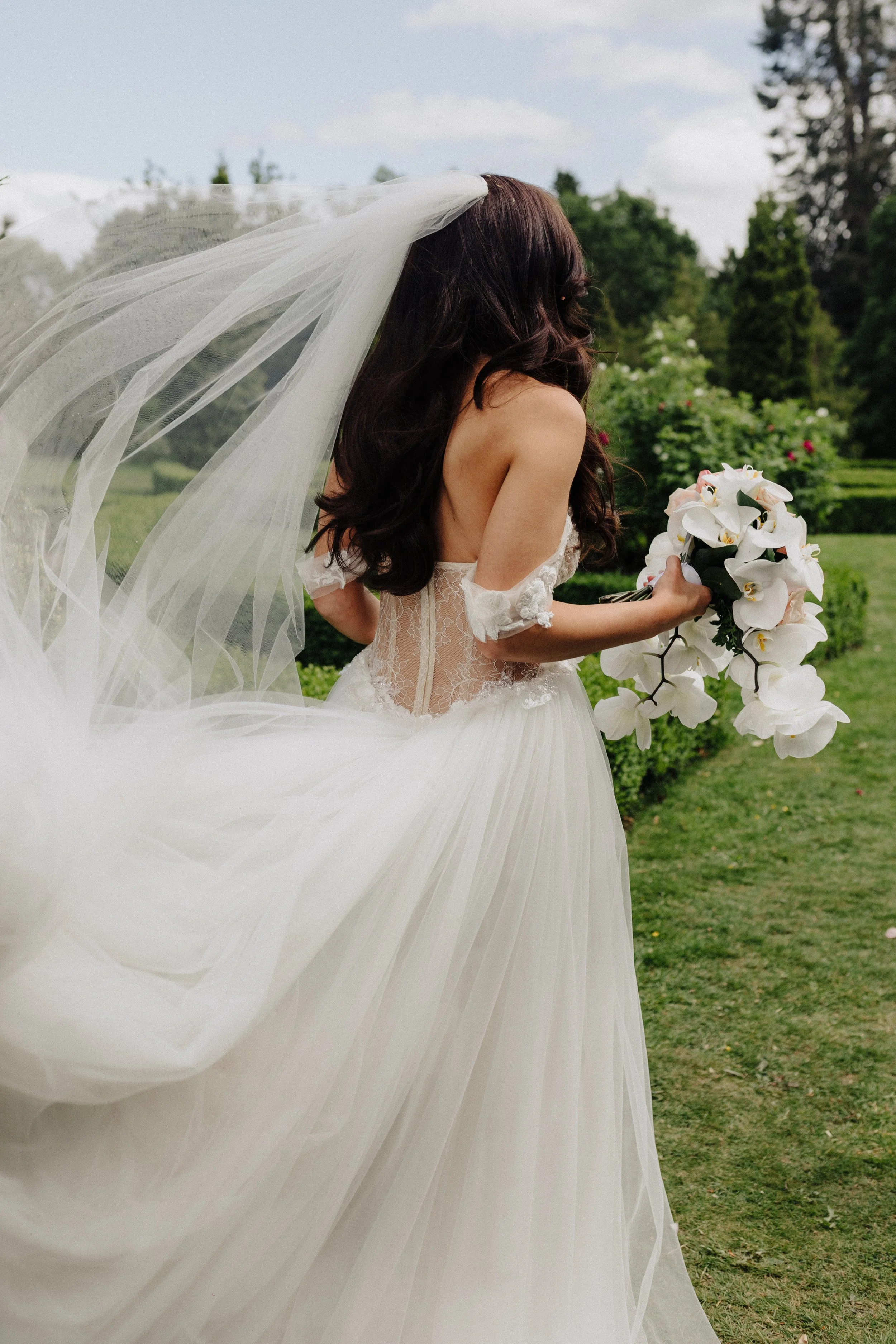 A bride in a white wedding dress with lace details, holding a bouquet of white orchids, outdoors in a green garden with trees and bushes, under a partly cloudy sky, with her long dark hair styled in loose curls and a wedding veil flowing behind her.