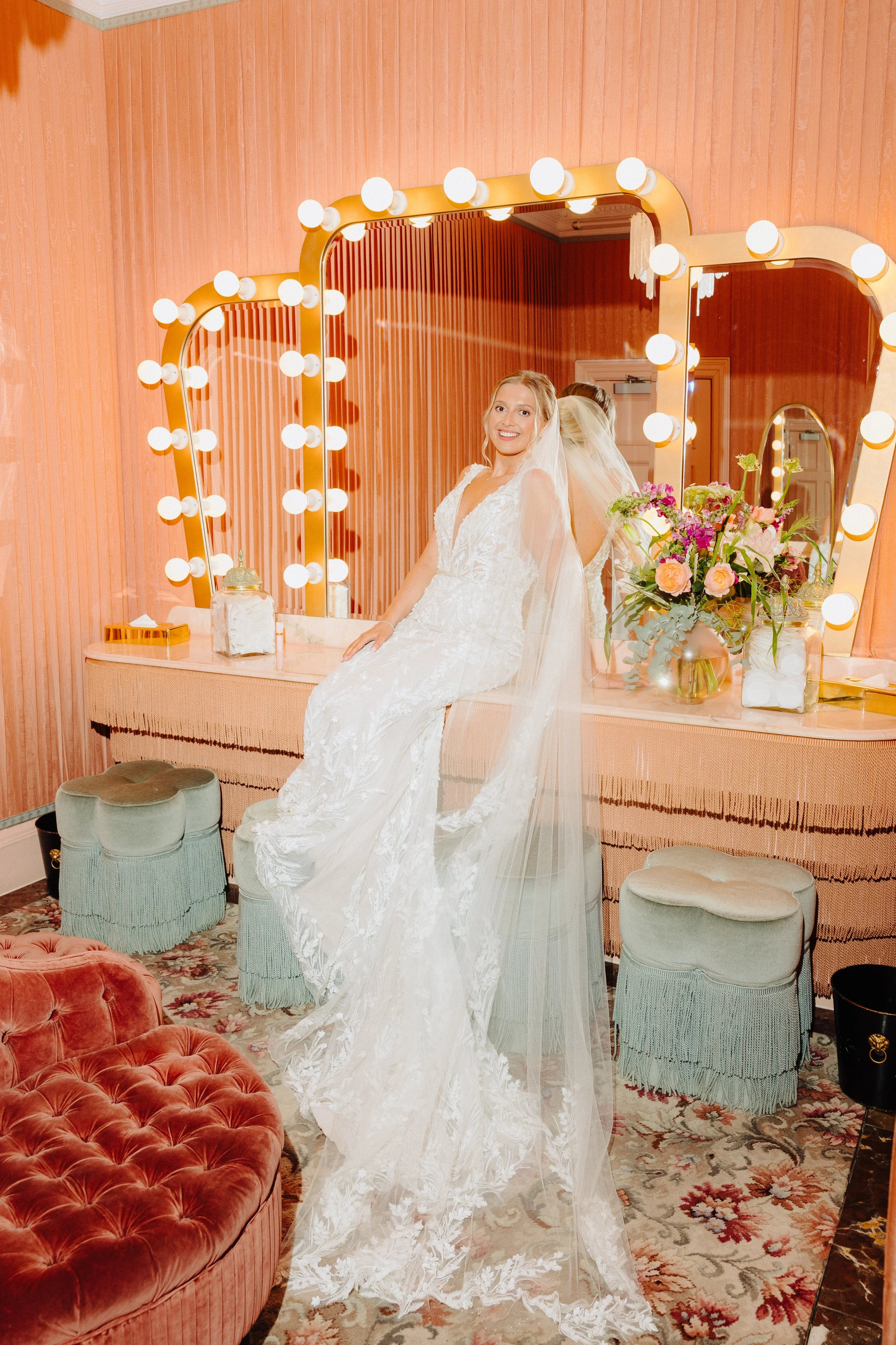 A bride in a white wedding dress and veil sitting at makeup vanity with illuminated mirror, flowers, and vintage decor in a pink-themed room.