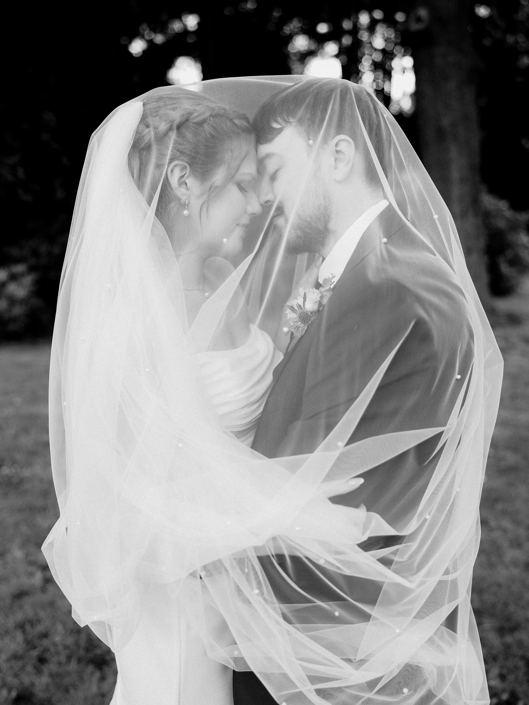 A black and white photo of a bride and groom with their foreheads touching beneath a veil, outdoors with trees in the background.