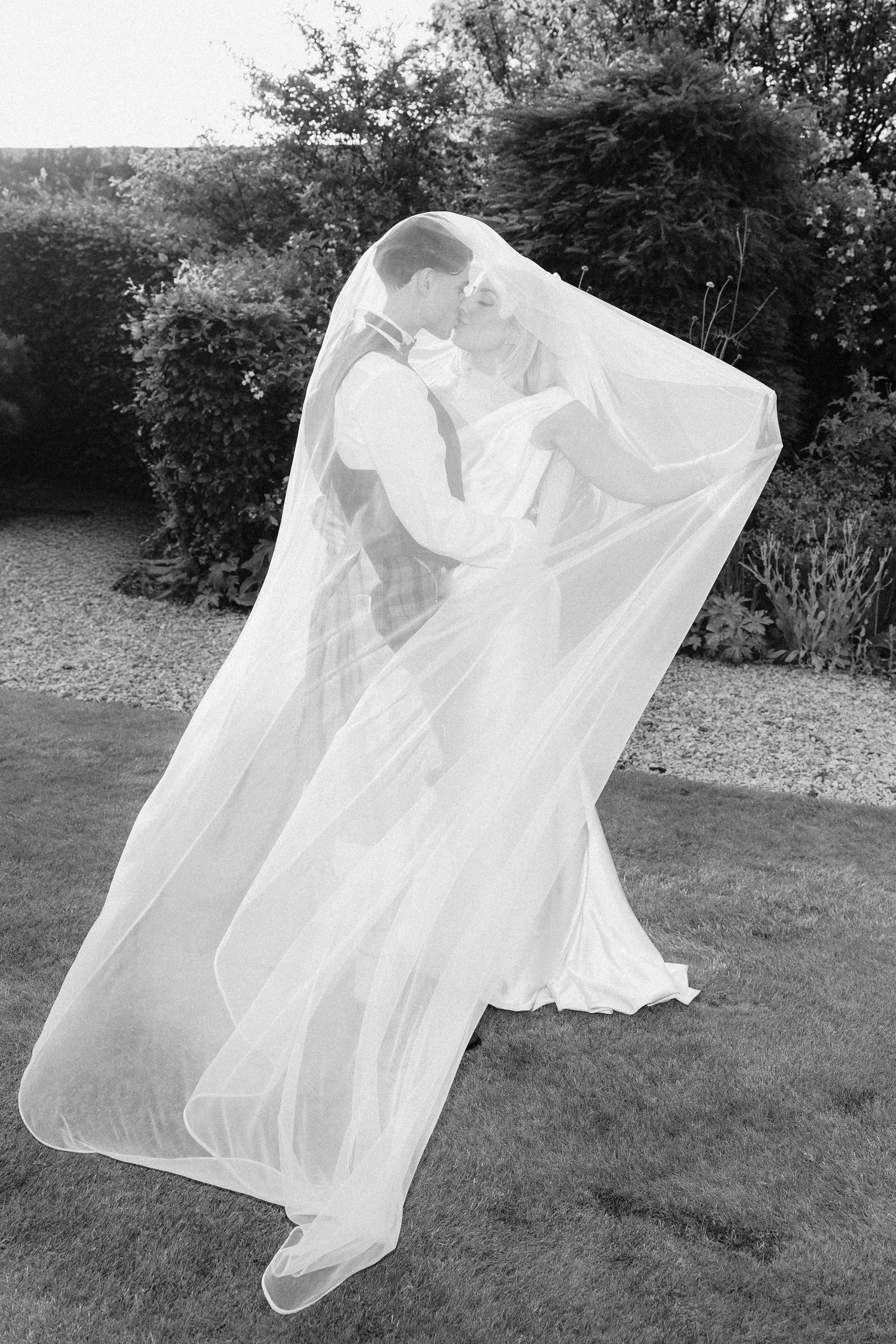 A bride and groom sharing a kiss outdoors with trees and bushes in the background. The bride is wearing a wedding dress and veil, and the groom is dressed in a suit. The bride's veil is flowing and she is holding onto the groom.