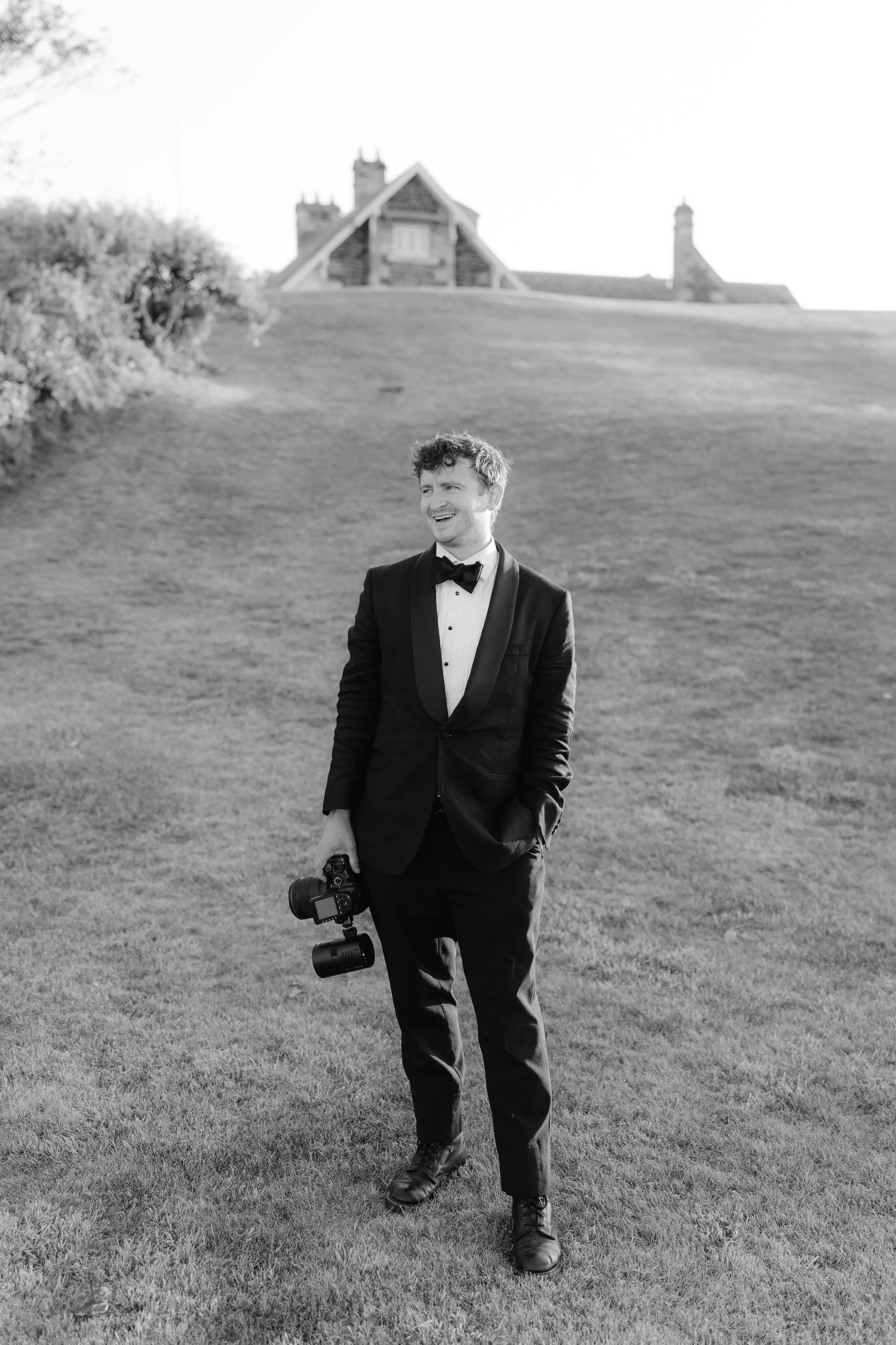 A man in a tuxedo holding a camera walks across a grassy field, with a large house in the background.