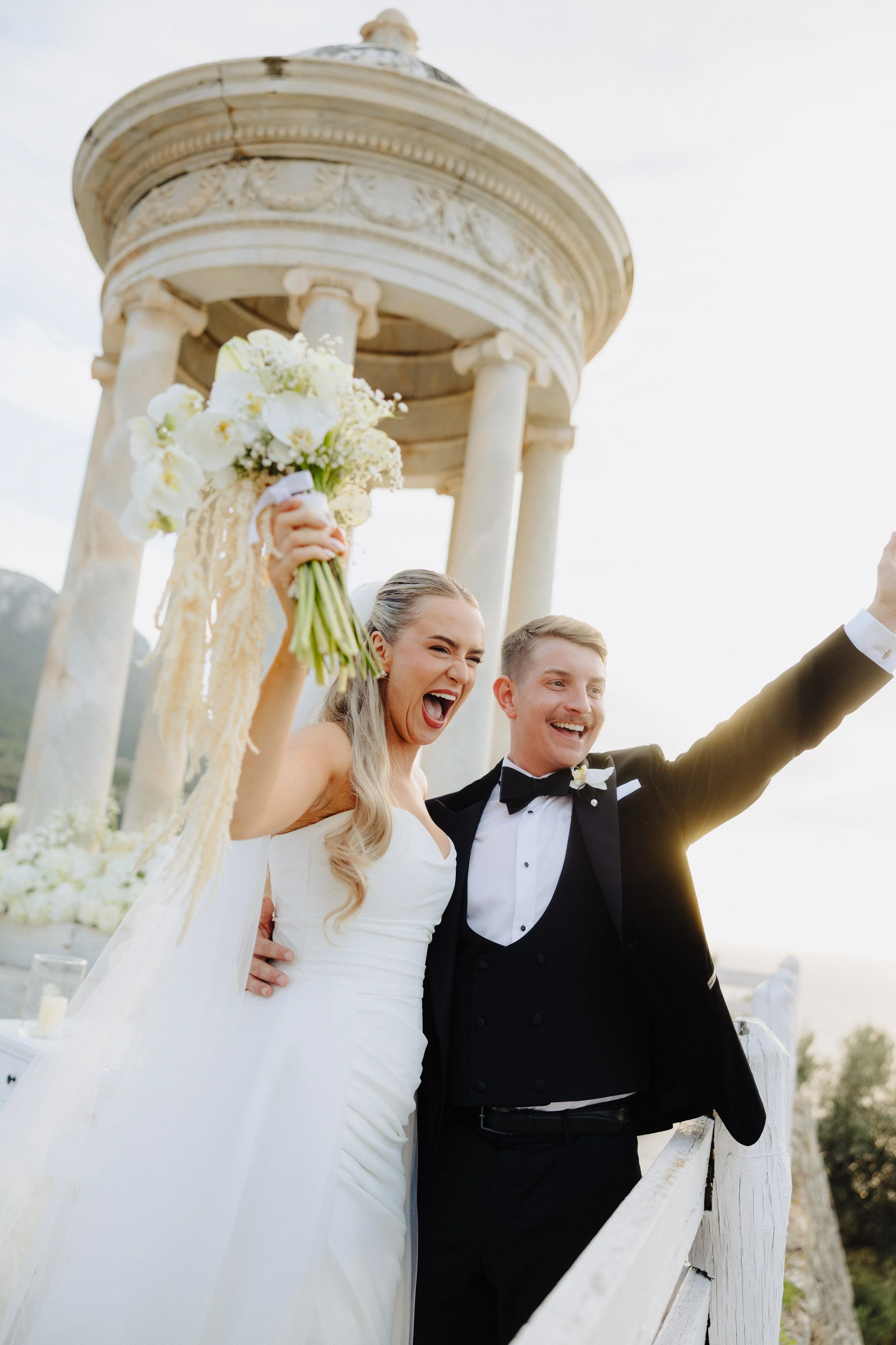 A smiling bride and groom celebrating on a bridge, with the bride holding a bouquet of white flowers and the groom wearing a black tuxedo, in an outdoor setting with a white classic-style pavilion in the background.