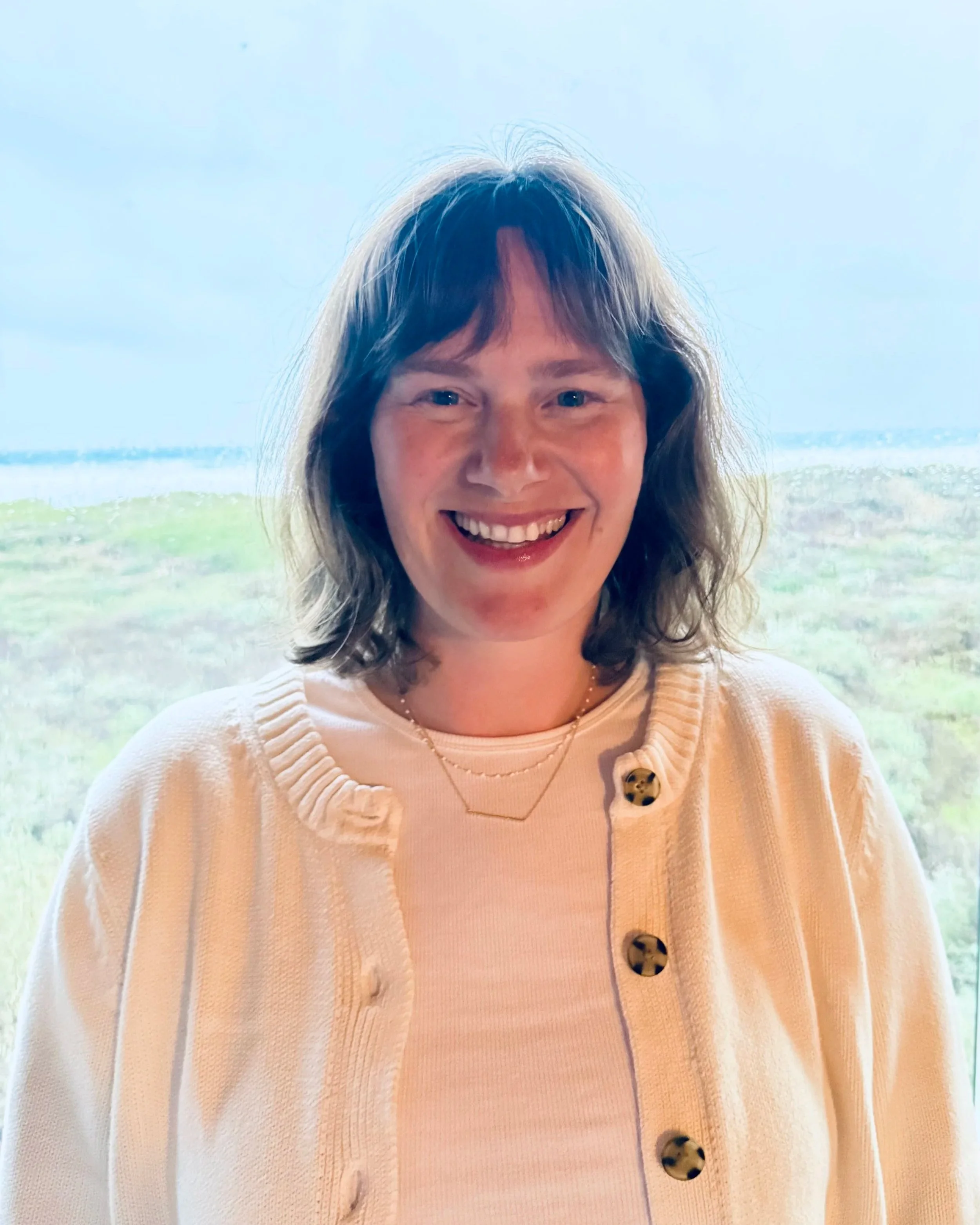 A smiling woman with shoulder-length brown hair wearing a beige cardigan with buttons, a light-colored top, and a delicate necklace, standing outdoors with a park or natural landscape in the background.