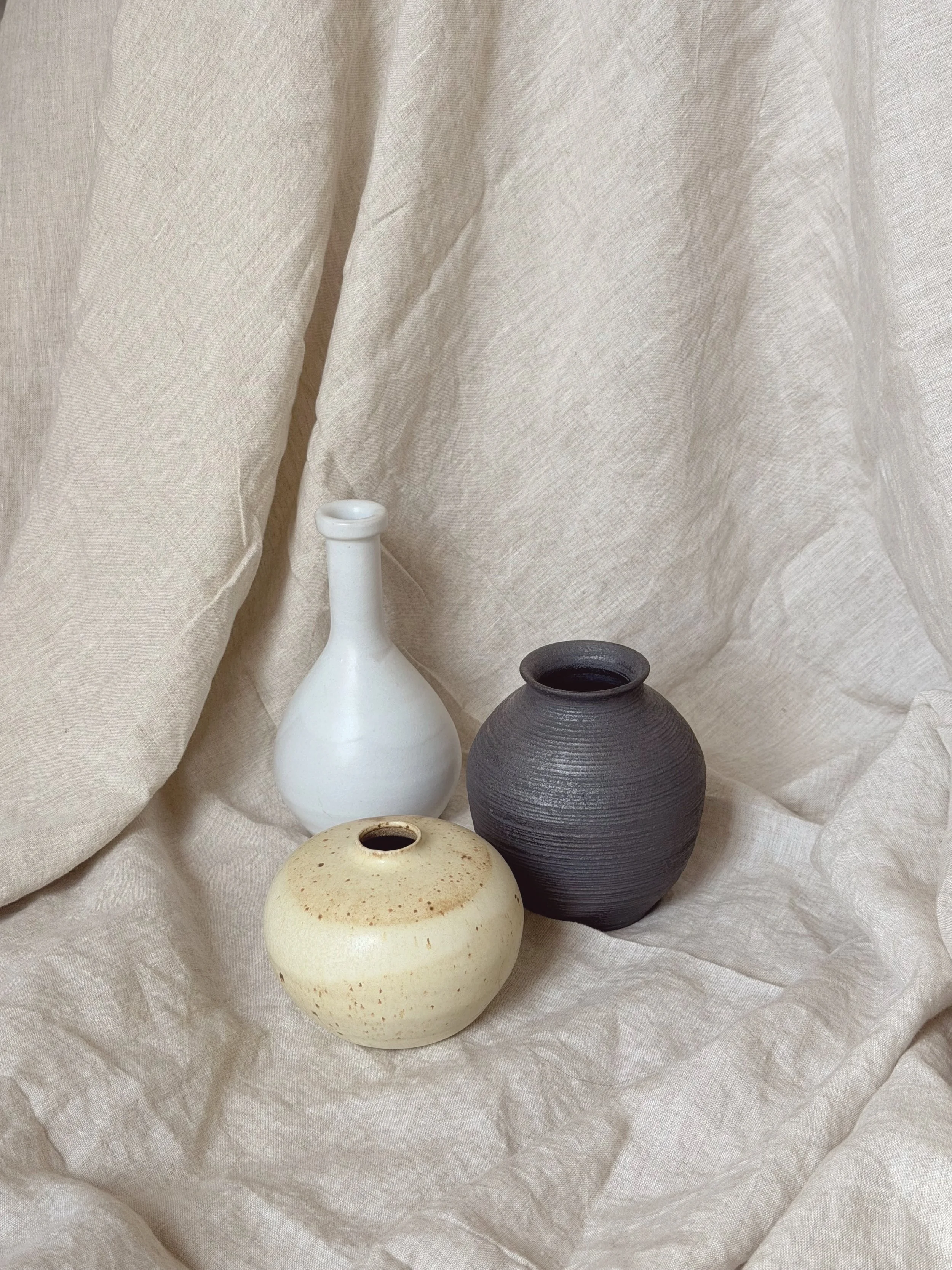 Three ceramic vases of different shapes and colors arranged on a fabric backdrop. The vases include a tall white one, a round beige one with speckles, and a dark gray one with a textured surface.