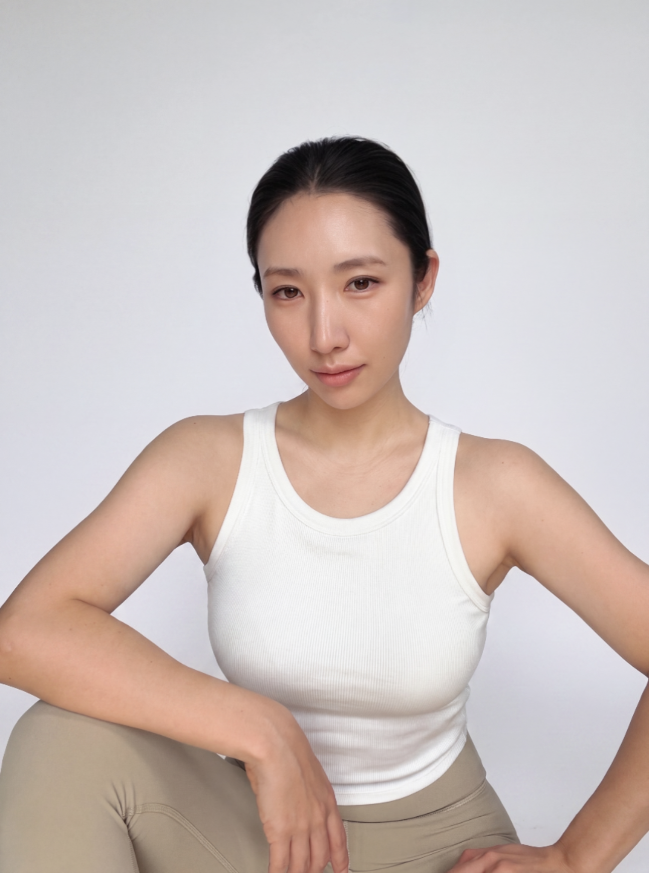 A woman with short black hair wearing a white tank top and beige leggings posing against a plain white background.