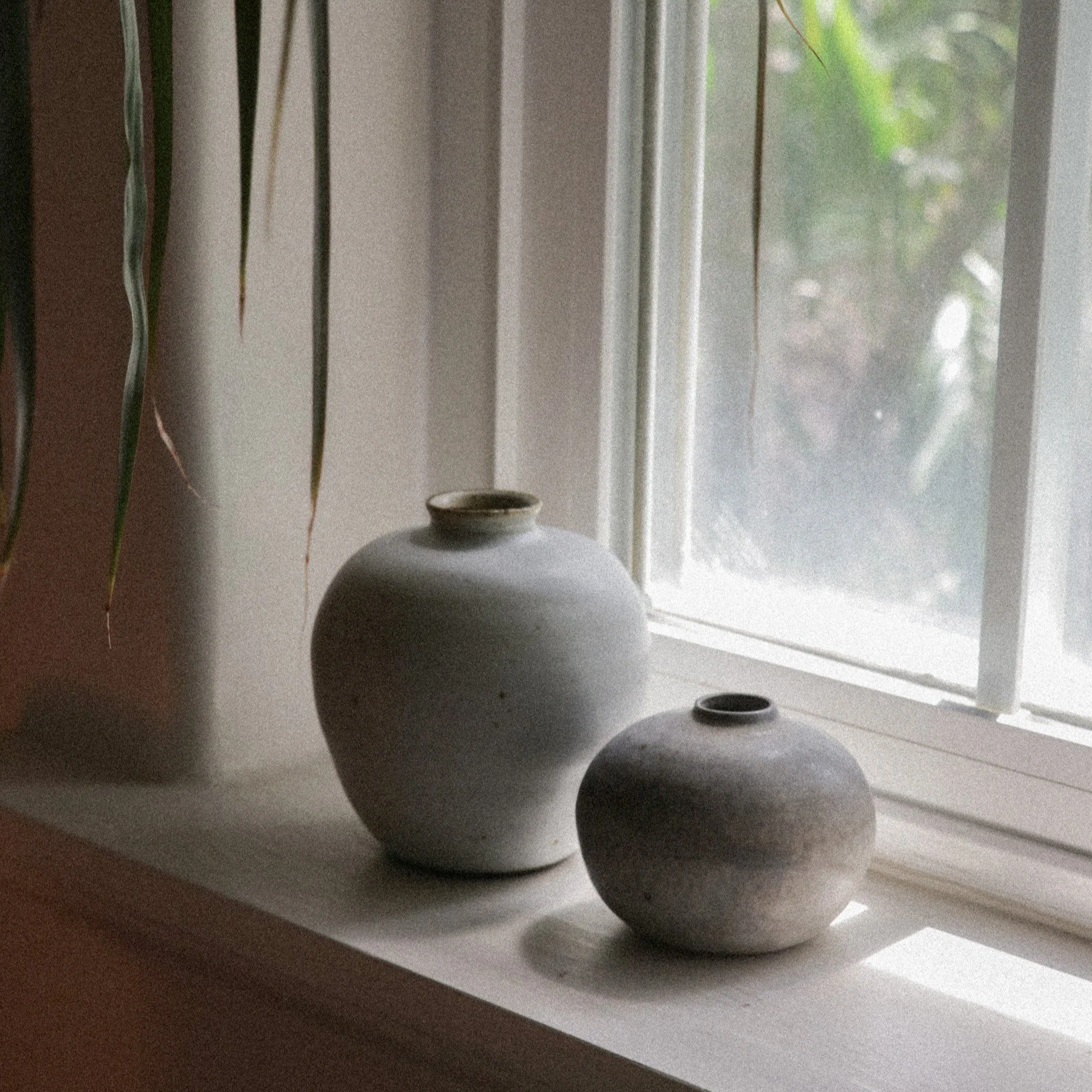 White ceramic vases on a windowsill, with a plant partially visible on the left and greenery outside through the window.