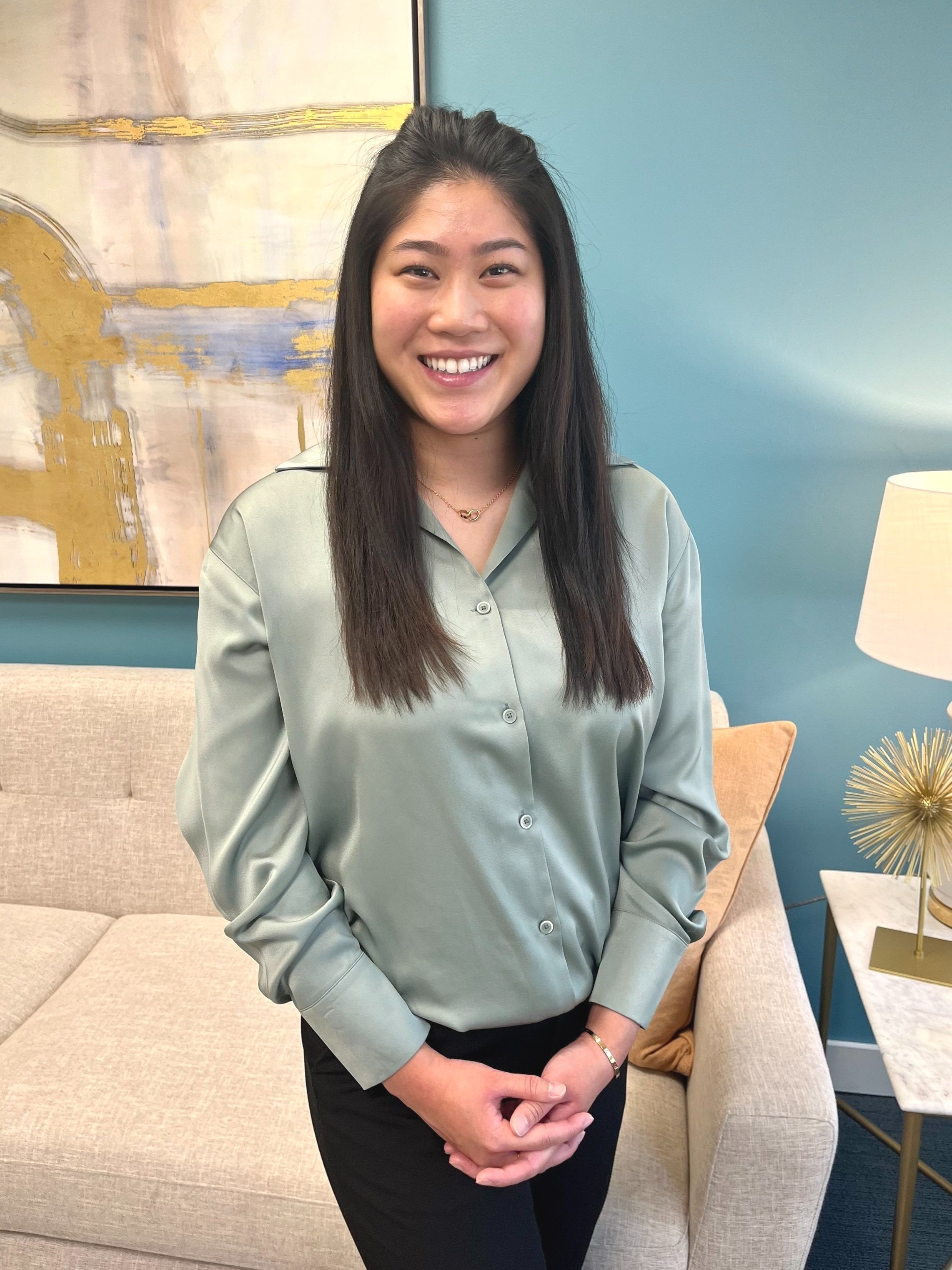Smiling woman in a light green blouse standing in a modern office with abstract art and a beige sofa.