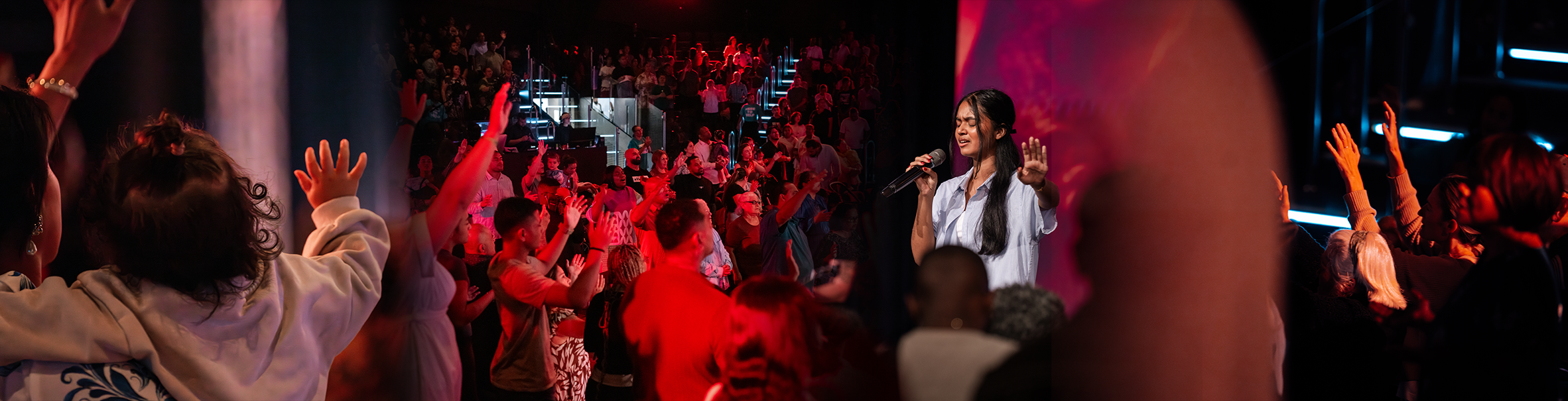 A large crowd with raised hands at a lively event. A woman with a microphone appears to be speaking or singing, engaging the audience. The scene is lit with red and pink lighting.