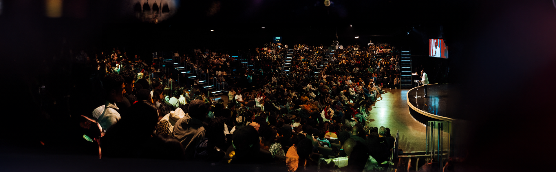 Large audience in a dimly lit auditorium listening to a speaker on stage.