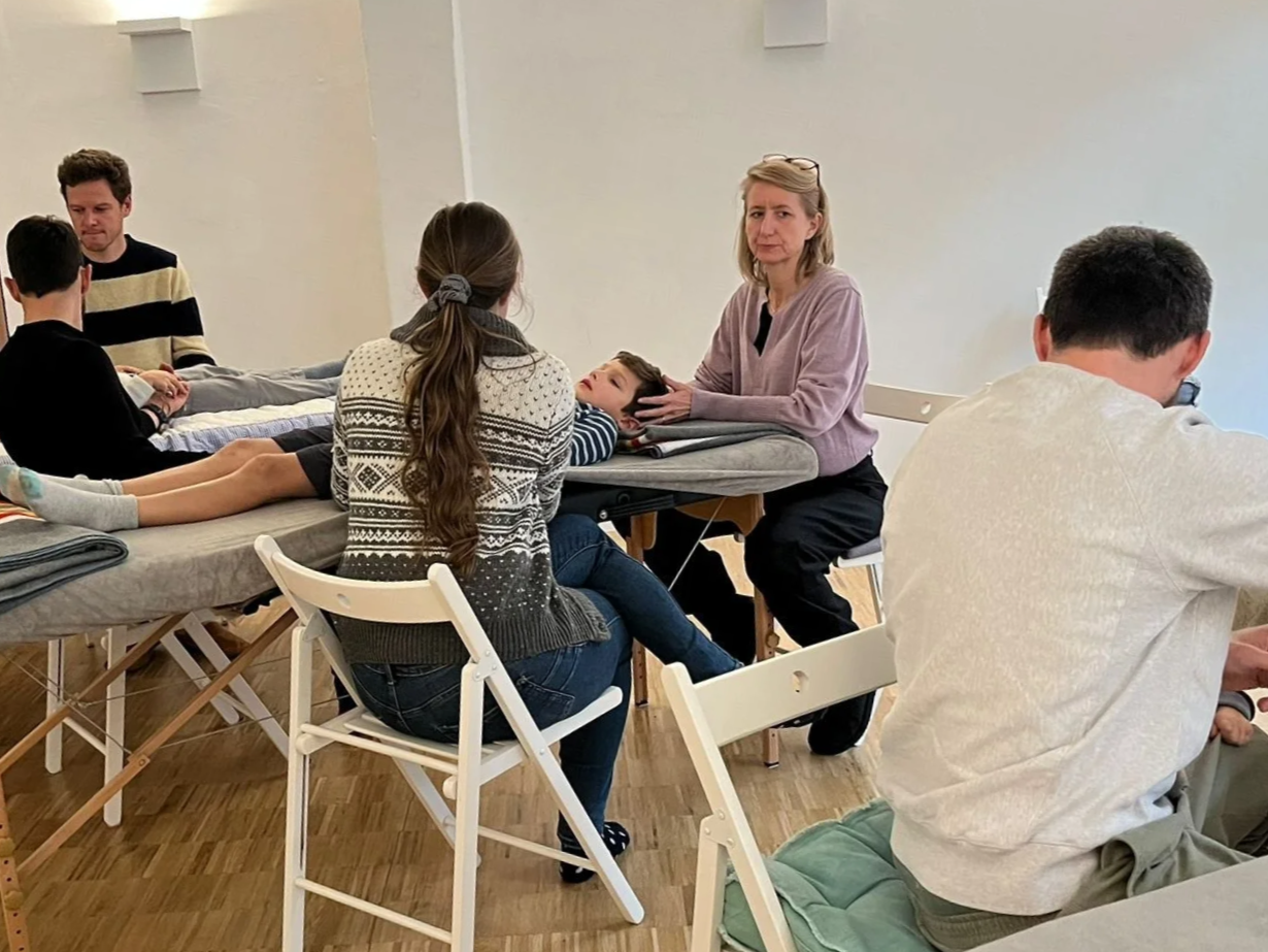 Une séance d'ostéopathie avec plusieurs personnes, un enfant allongé sur une table d'examen, et un ostéopathe parlant à un adulte. La scène se déroule dans une pièce claire avec un sol en bois.