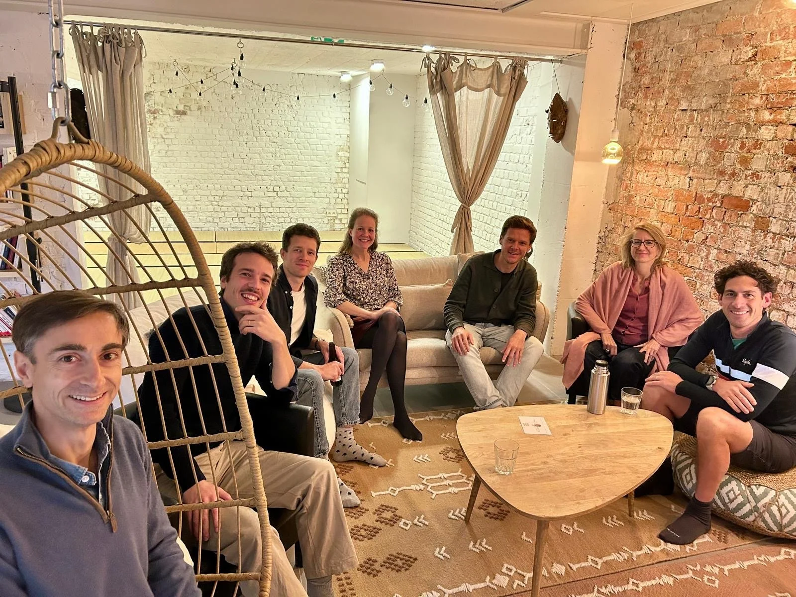 Groupe de sept personnes assises dans un salon, souriantes, avec un mur en briques et une décoration chaleureuse.