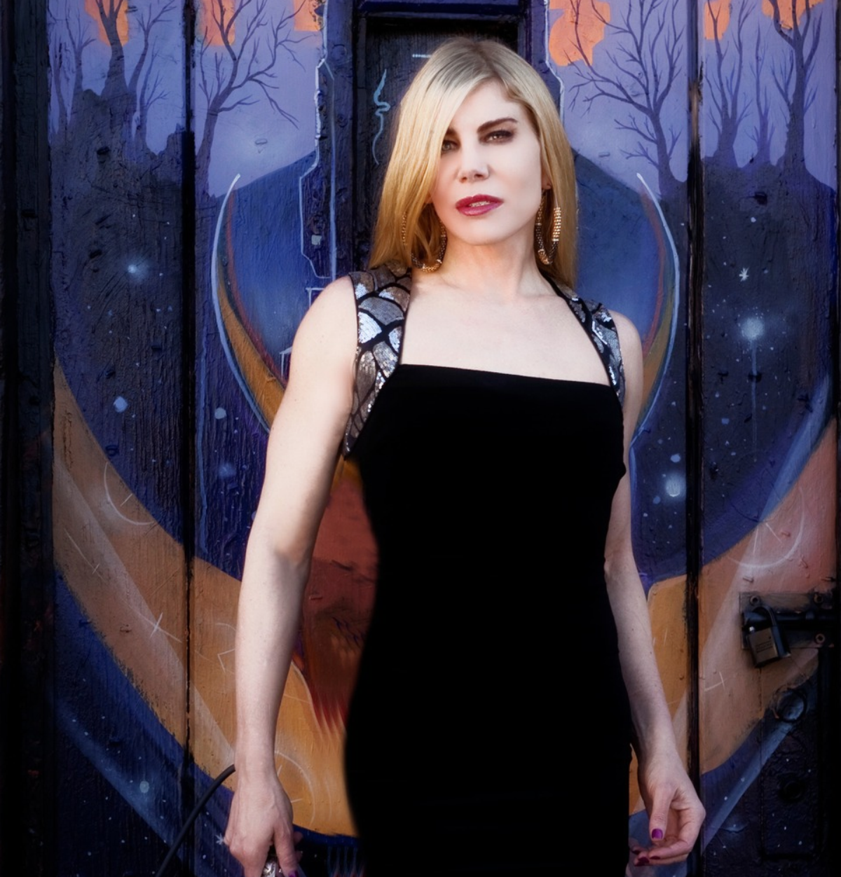 Portrait of singer-songwriter Meghan Monroe in a black gown, standing before a colorful painted mural featuring blue and violet architectural details, silhouetted trees, and a warm rose-colored sky.