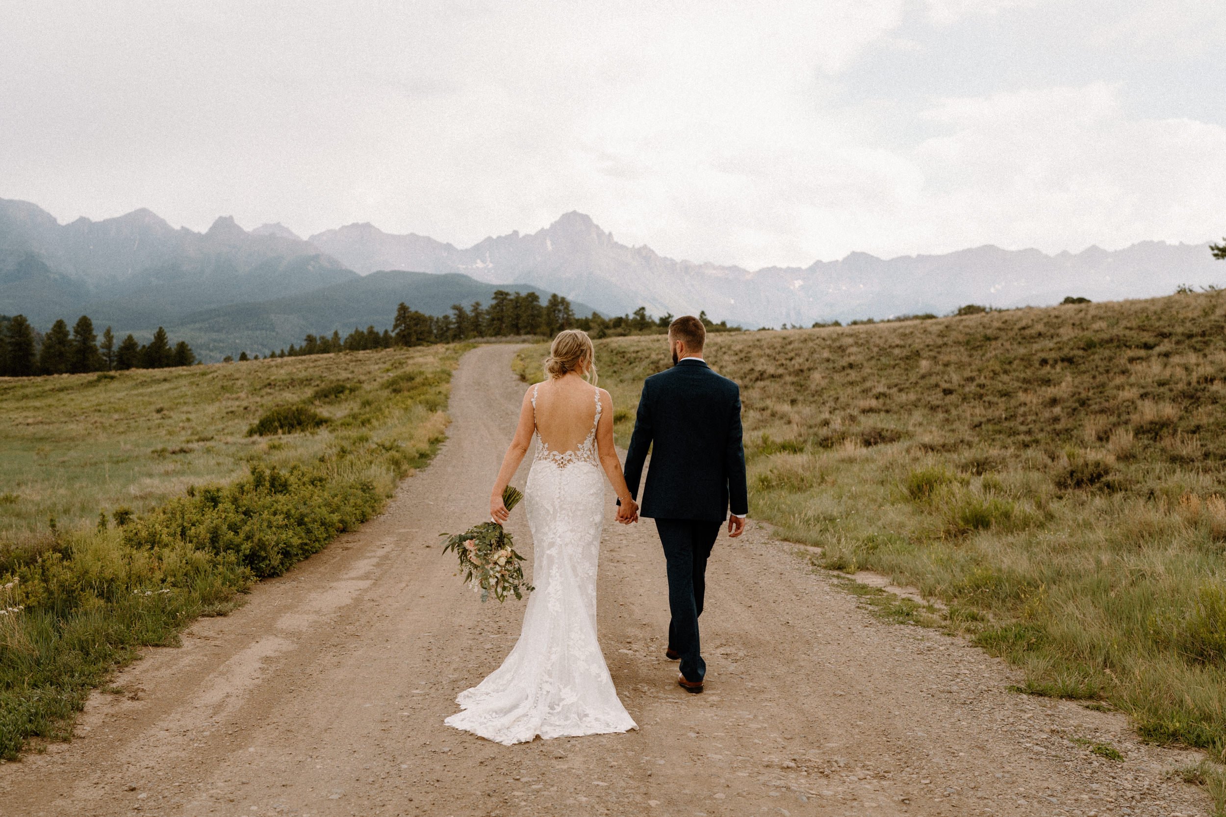 Bride and groom hold hands and walk down dirt road at Top of the Pines in Ridgway, CO