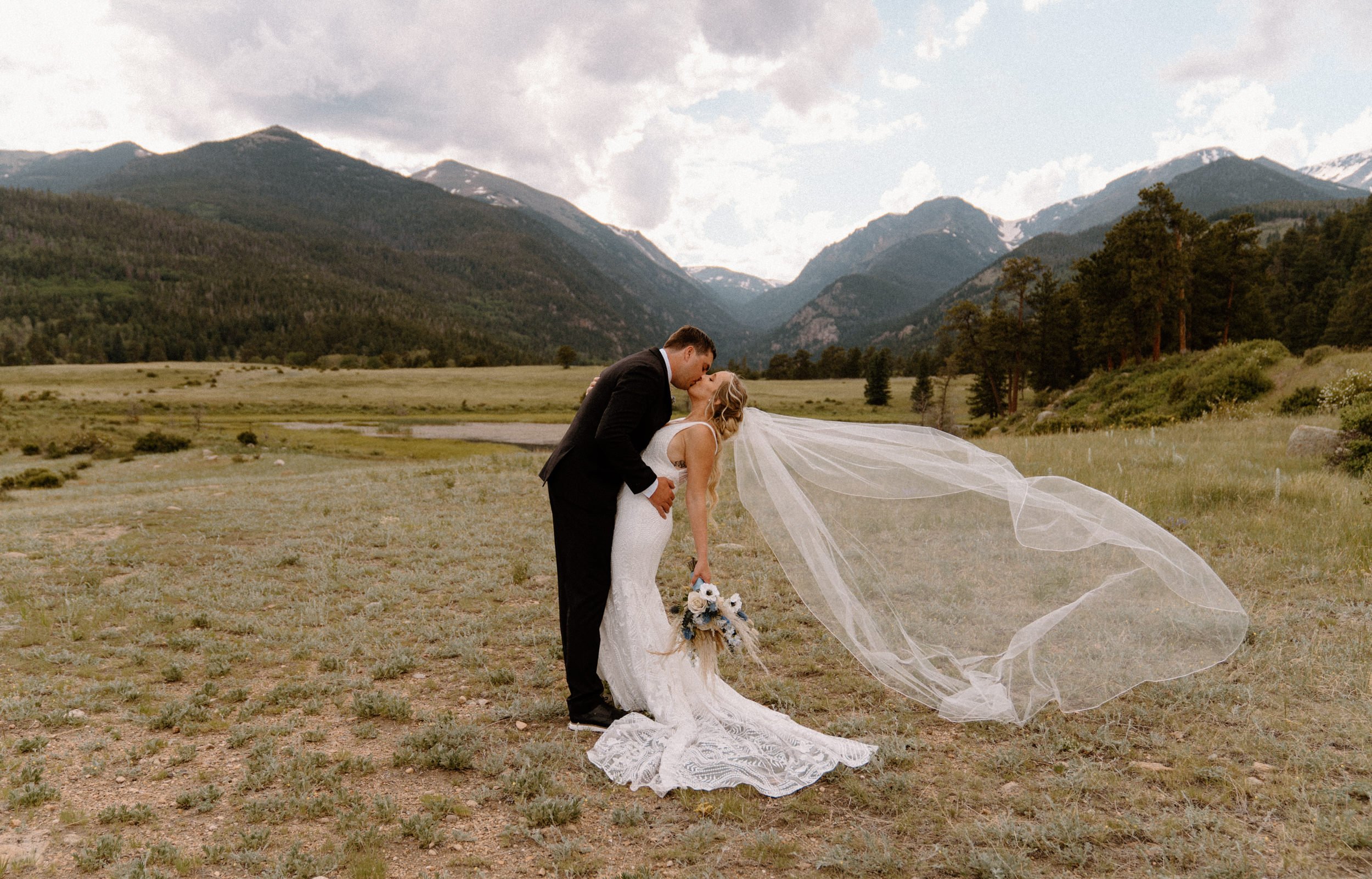The bride and groom kiss in a mountain valley as her veil blows out behind her