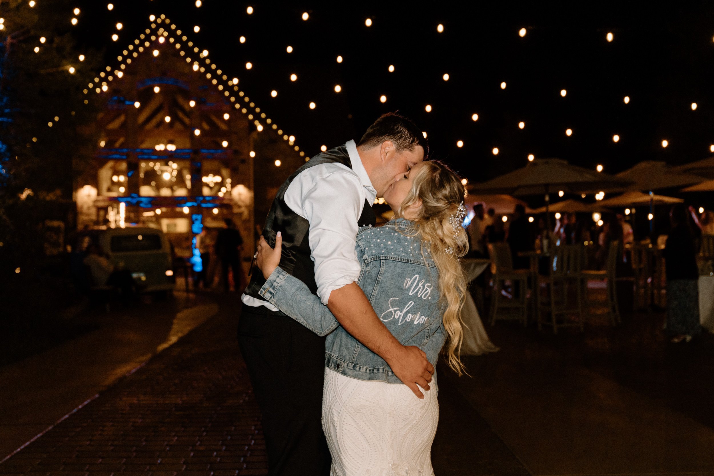 The bride and groom kiss; the bride is wearing a jean jacket that says "Mrs. Solomon" on the back