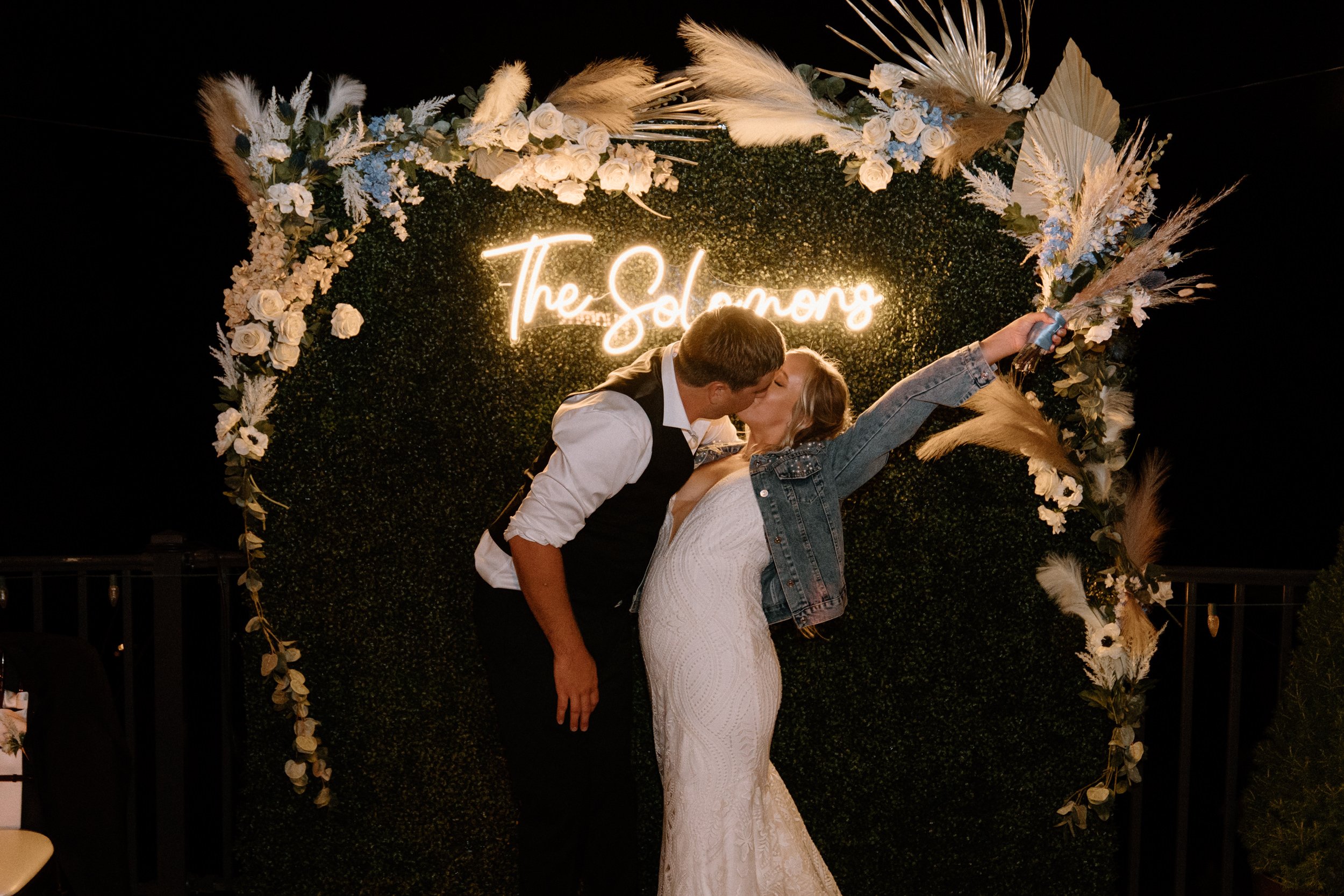 The bride and groom kiss in front of the neon sign that reads "The Solomons"