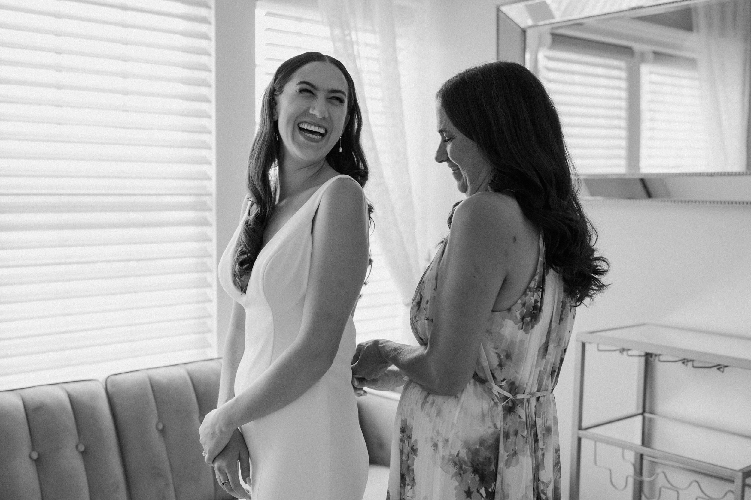 Bride smiles back at a bridesmaids as she helps her into the wedding dress