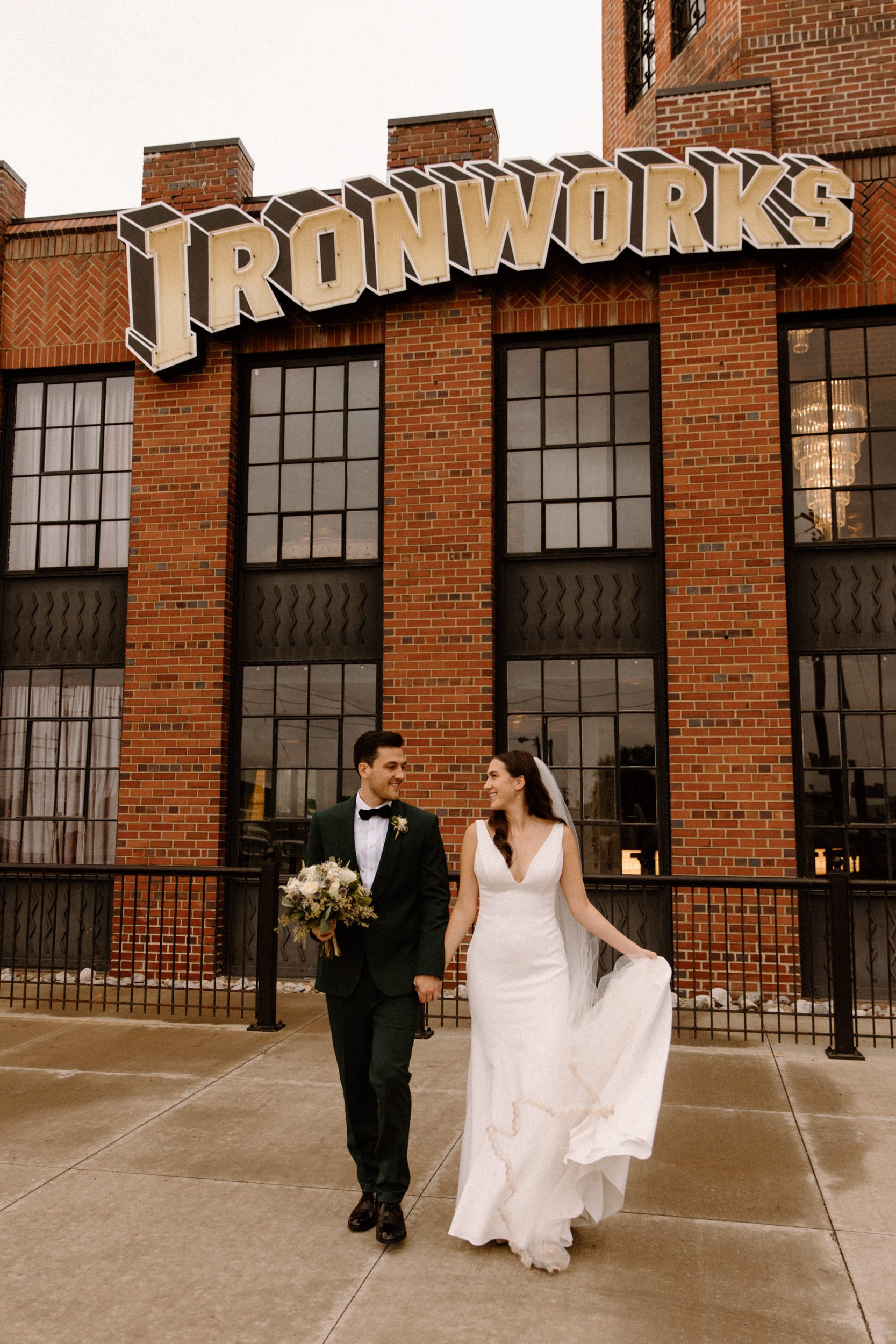 The bride and groom pose under the Ironworks sign in front of a brick building