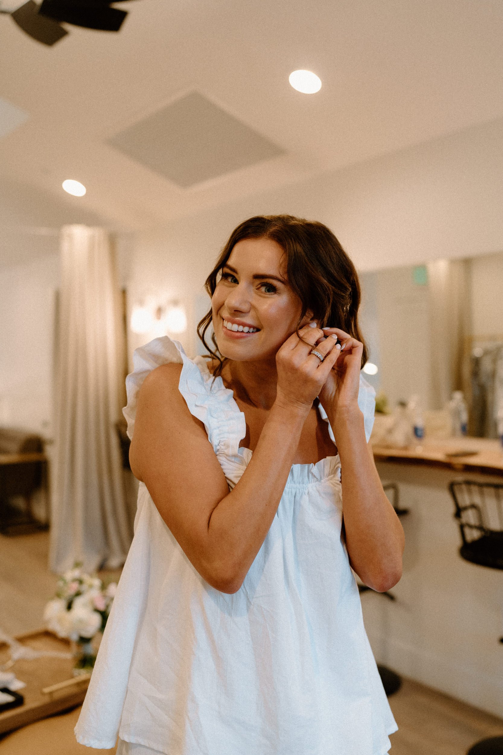 The bride smiles at the camera as she puts an earring in her ear
