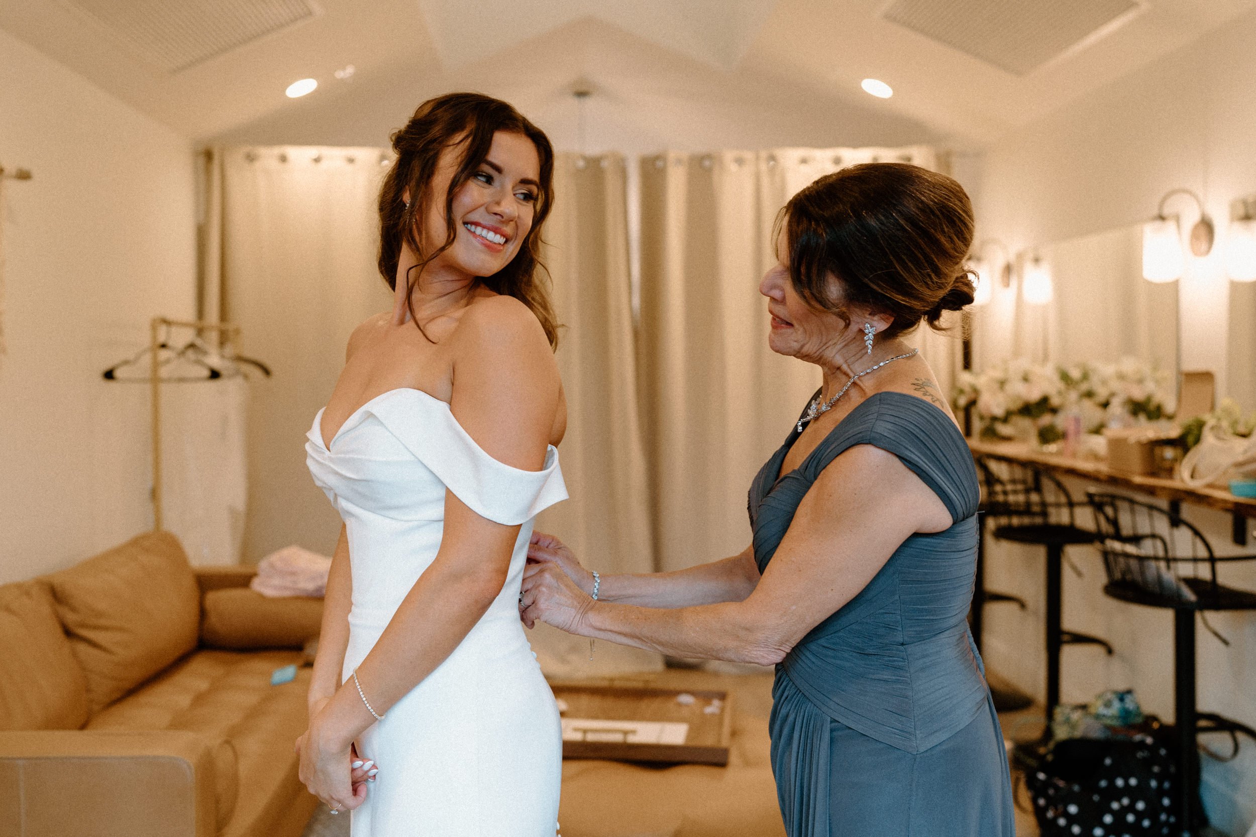The bride smiles back at her mother as she helps button her wedding dress