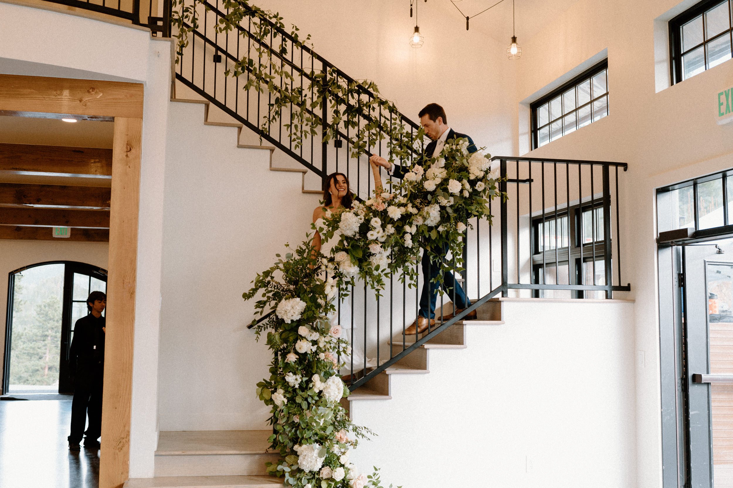 The bride and groom walk down the stairs decorated with greenery and white roses