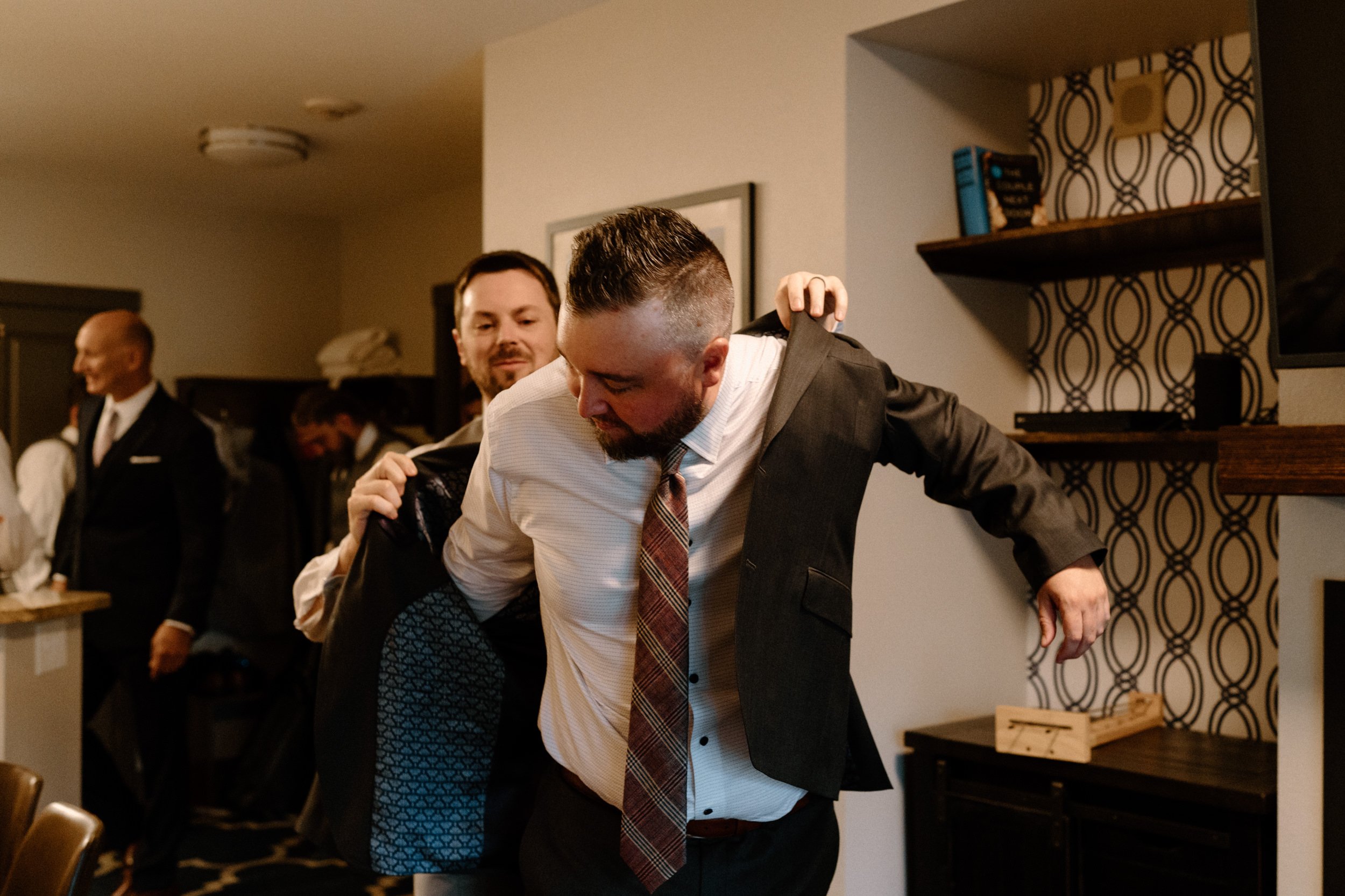 A groomsmen helps the groom into his suit jacket