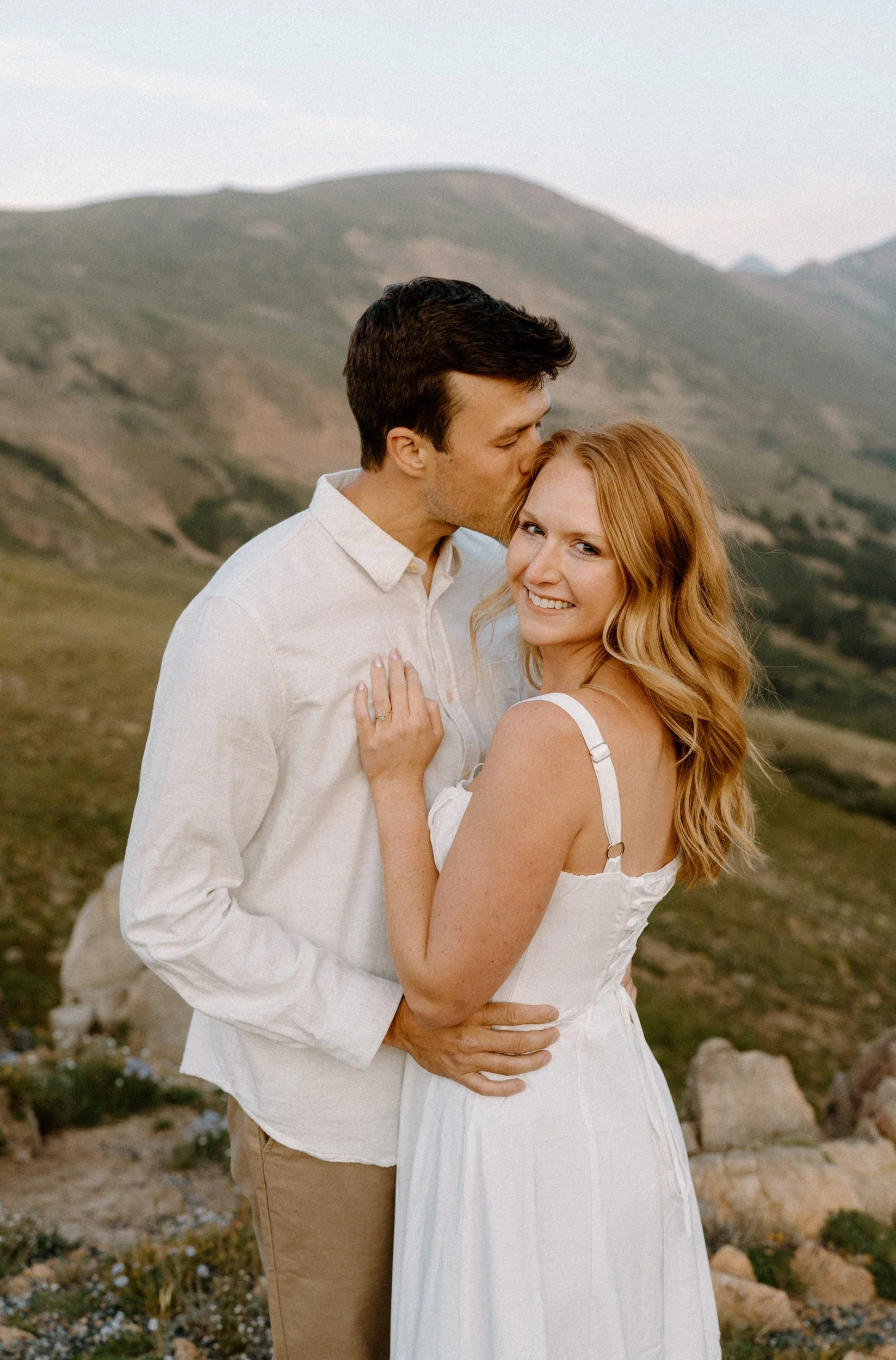 Fiancée smiling at camera while fiancée kisses her forehead at Loveland Pass during engagement session