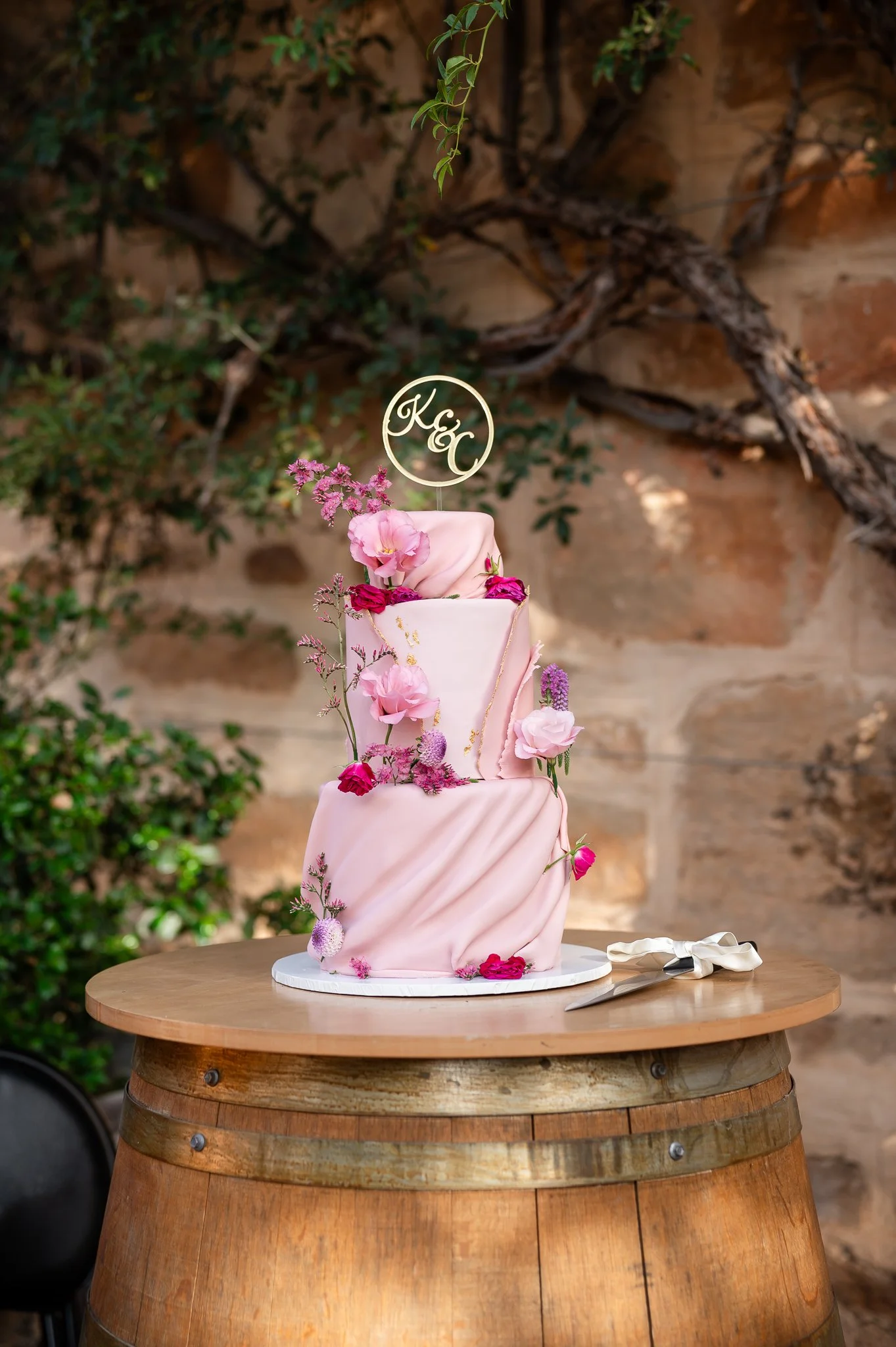 A multi-tiered pink and white wedding cake decorated with flowers sits on a round table in front of a rustic brick wall and greenery. A cake knife and server are placed beside it.
