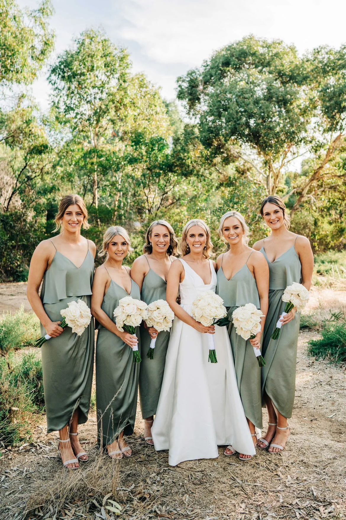 Group of six women dressed in formal attire, holding white bouquets, standing outdoors with trees in the background.
