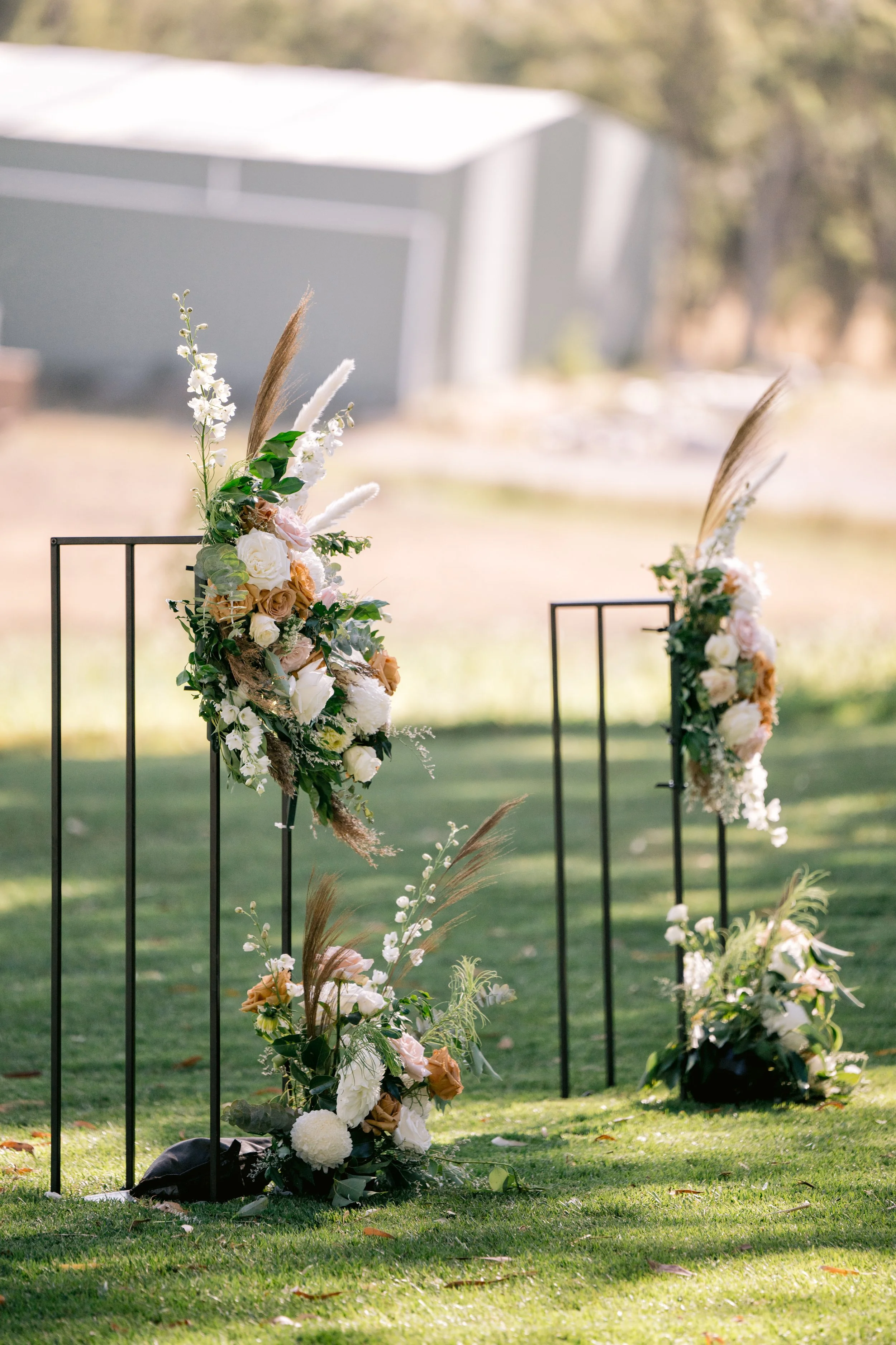 Decorative floral arrangement with white and peach roses, greenery, dried pampas grass, and other flowers on black metal stands outdoors during daytime.