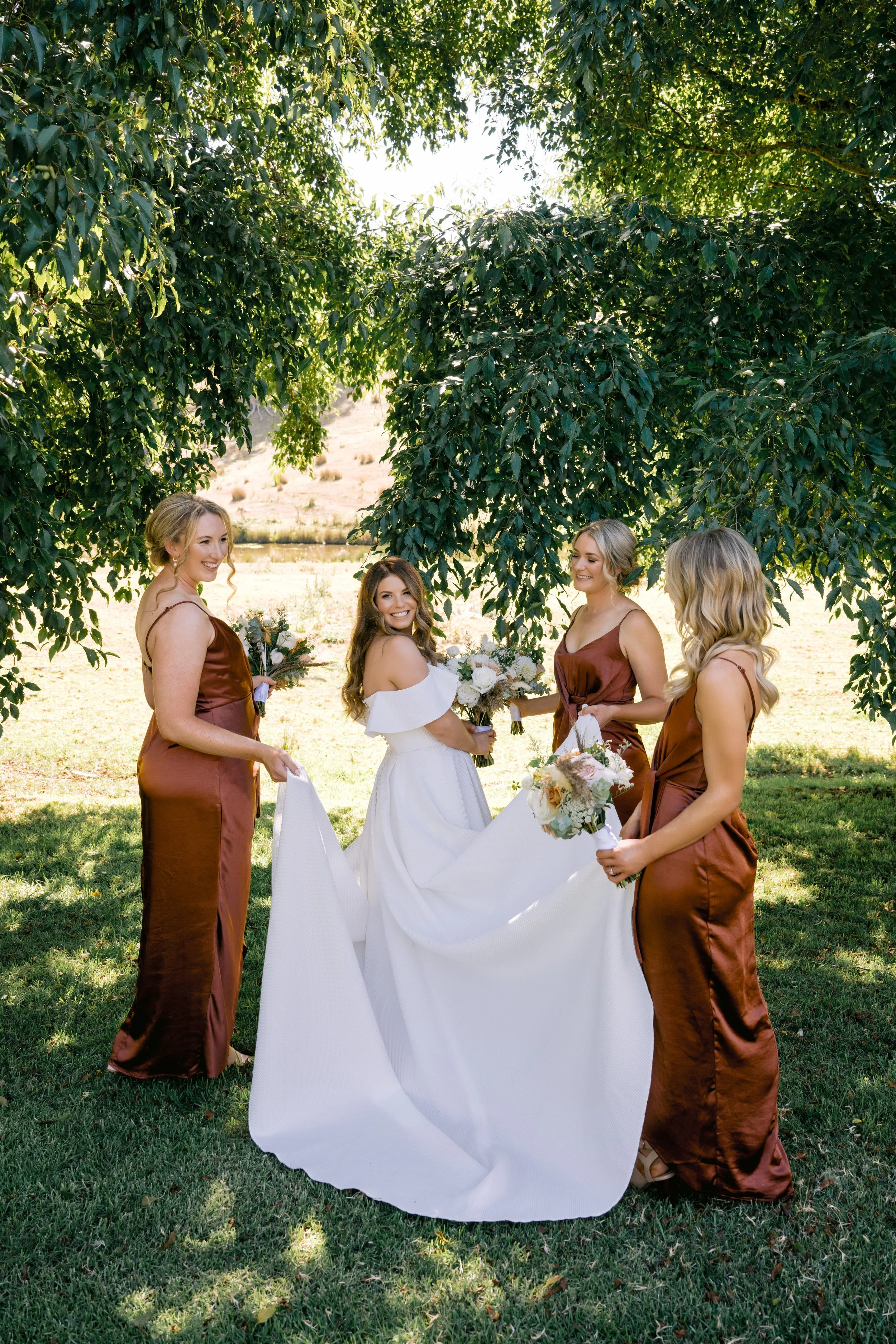 A bride and four bridesmaids standing outdoors under a large leafy tree. The bride is in a white wedding dress holding a bouquet, and the bridesmaids are in matching rust-colored satin dresses holding bouquets. They are smiling and looking at each other in a natural, sunny setting.