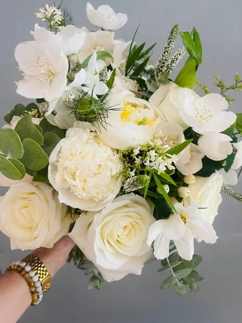 Close-up of a bouquet of white flowers, including roses, peonies, and other blooms, with green foliage and small white accents.