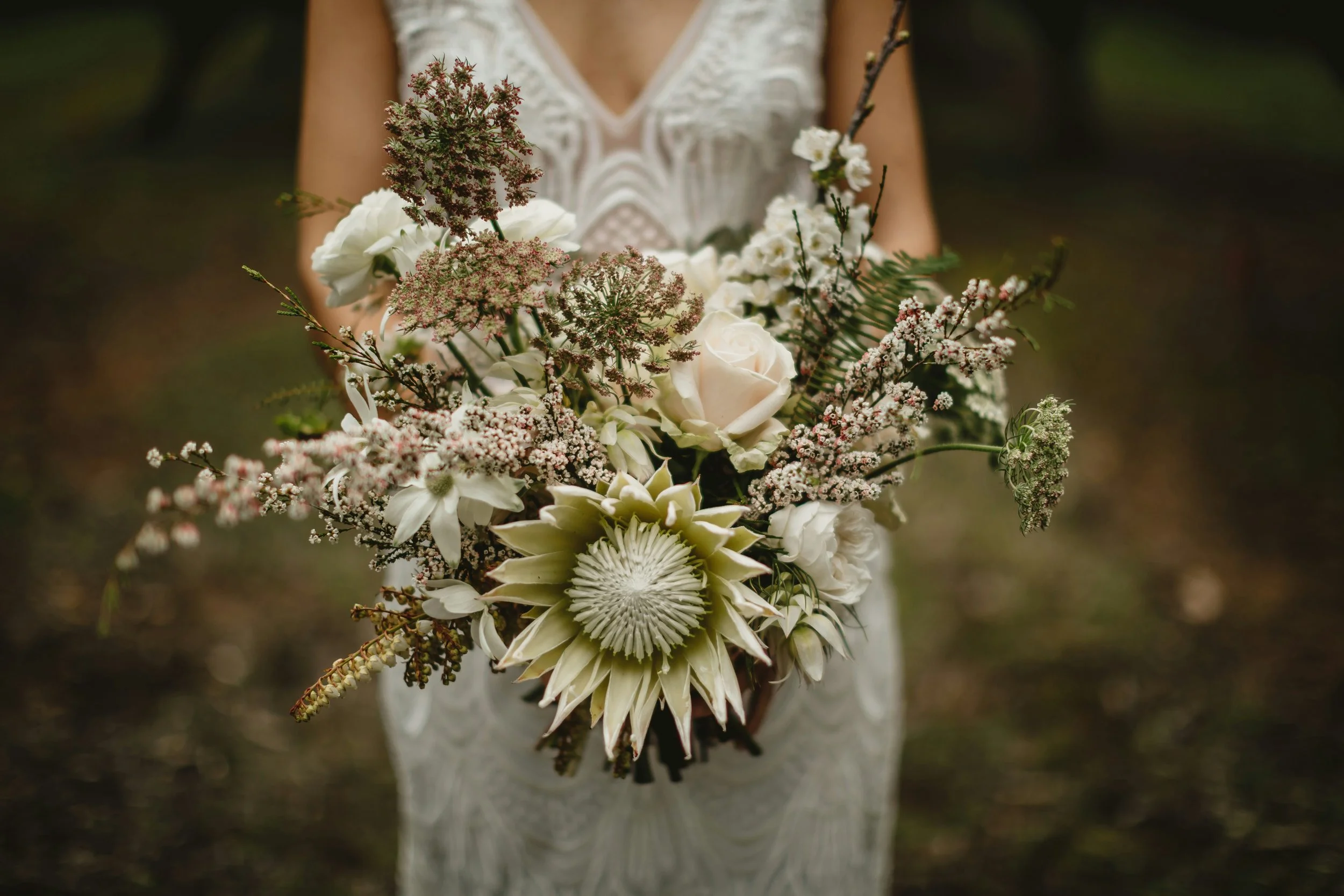 A woman in a white lace dress holding a bouquet of various white and pink flowers and greenery outdoors.