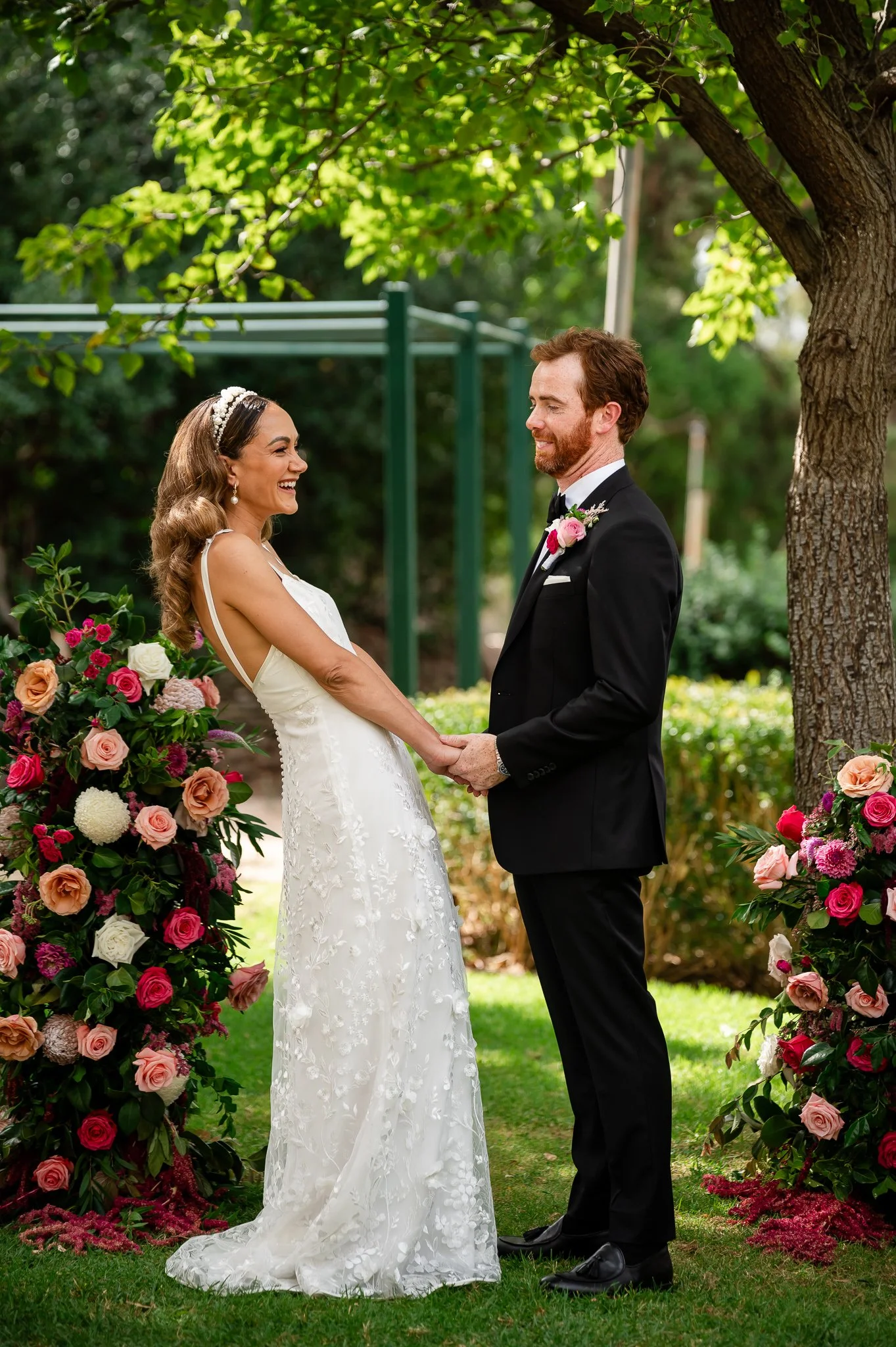 A bride and groom stand facing each other outdoors, holding hands, with wedding flowers and greenery around them.