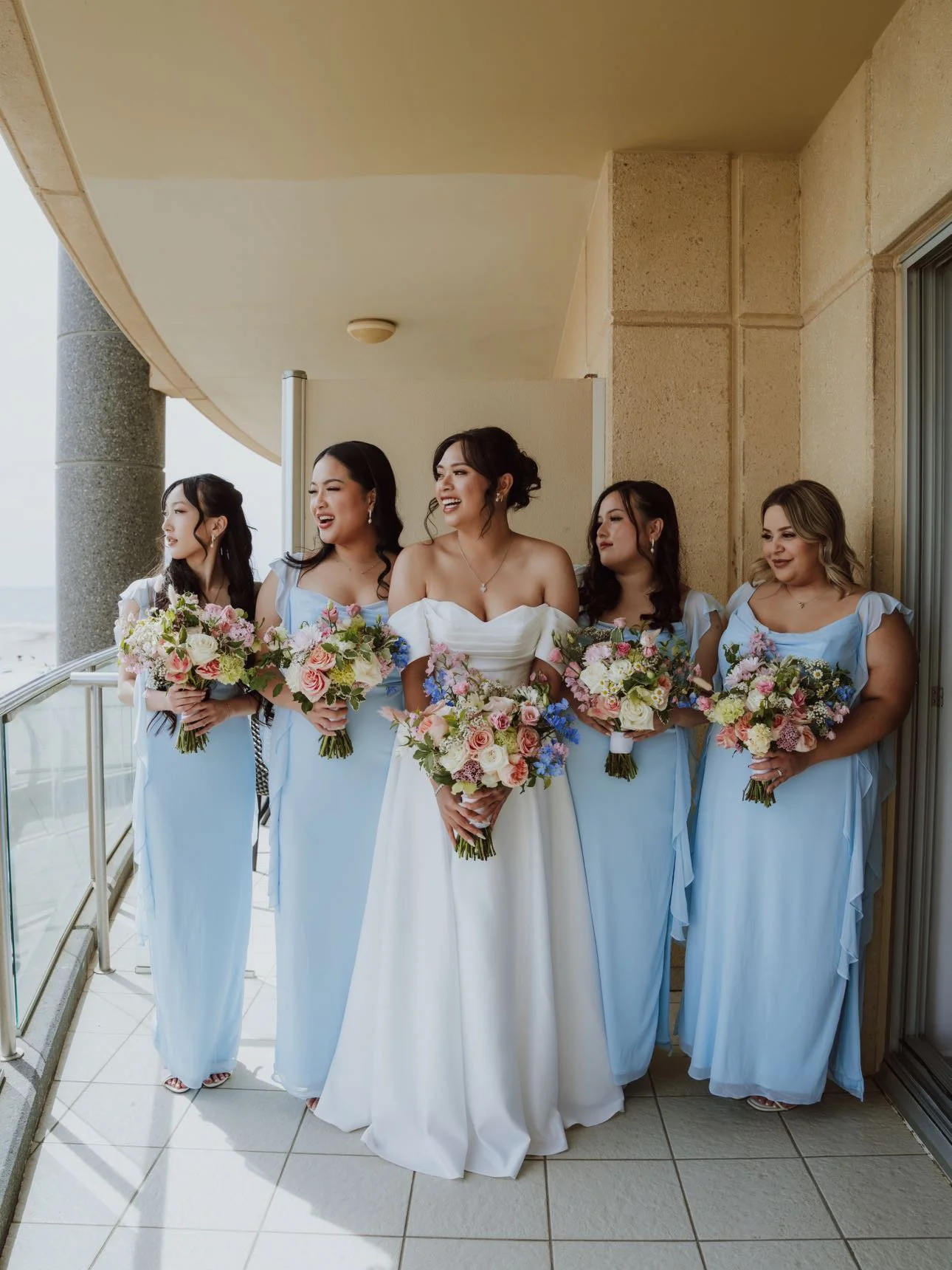 A bride and five bridesmaids on a balcony, all holding bouquets of flowers. The bride is in a white gown, and the bridesmaids are in light blue dresses. They are smiling and looking in different directions.