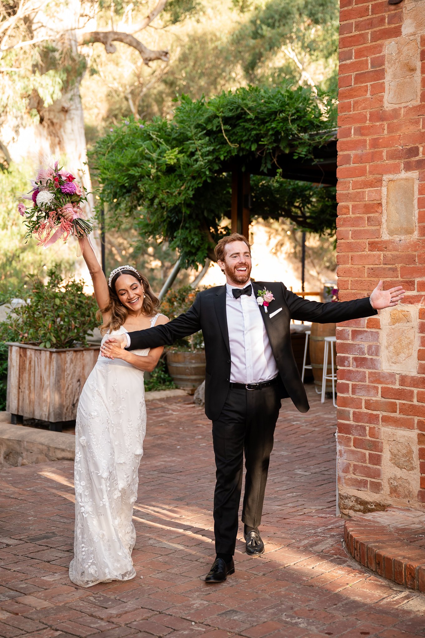A bride in a white wedding dress and a groom in a black tuxedo are joyfully celebrating outdoors. The bride holds a pink and purple bouquet in her right hand raised high and smiles happily. The groom extends his arms outward with a big smile. They walk on a brick pathway next to a brick wall, with sunlit trees and greenery in the background.