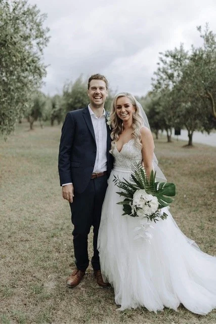 A newlywed couple standing outdoors, smiling, with trees in the background. The bride is holding a bouquet and wearing a white wedding dress, while the groom is dressed in a navy suit.