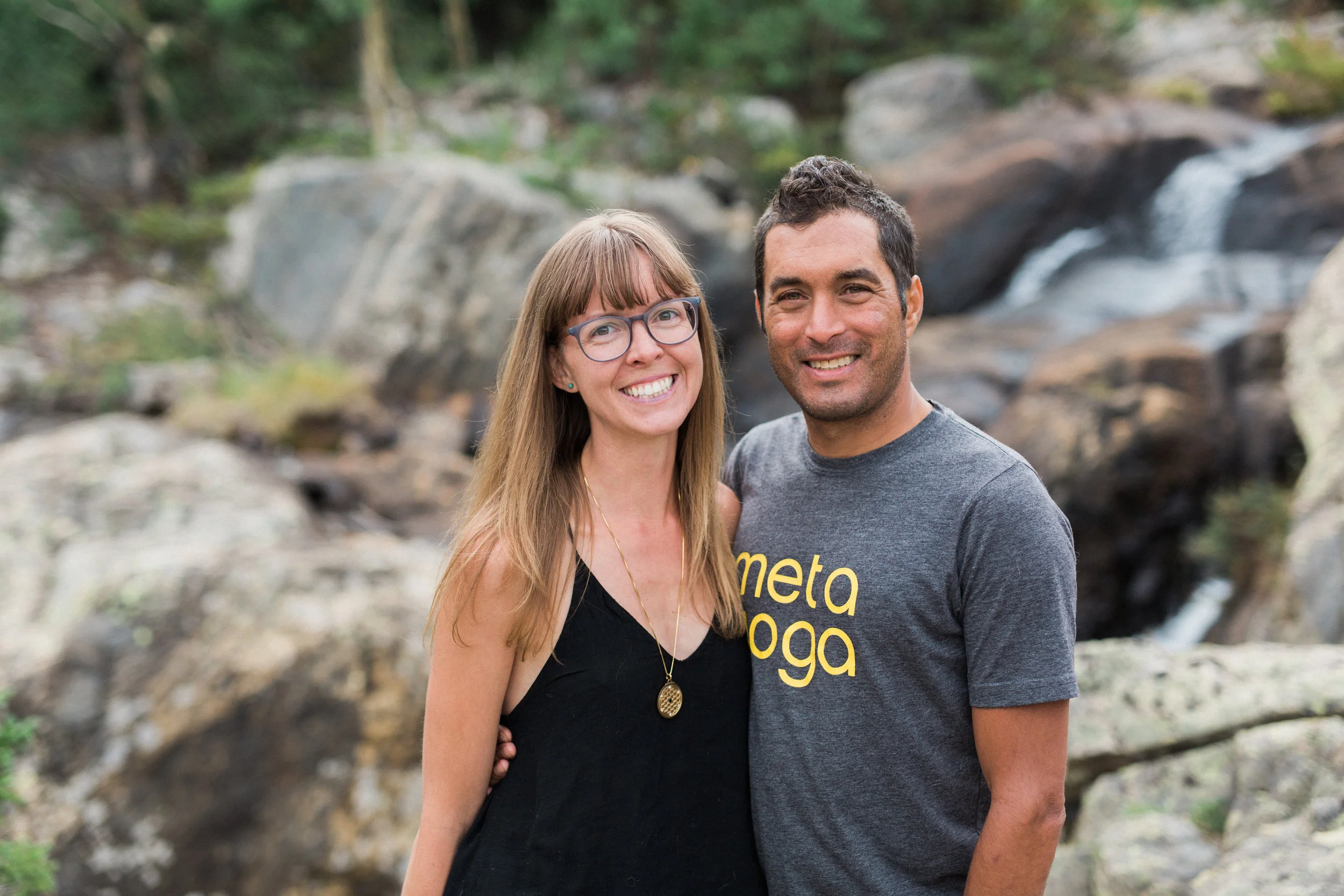 A smiling couple outdoors near a rocky stream, with trees and greenery in the background. The woman has long blond hair, glasses, and is wearing a black top and gold necklace. The man has short dark hair, and is wearing a gray T-shirt with yellow text.