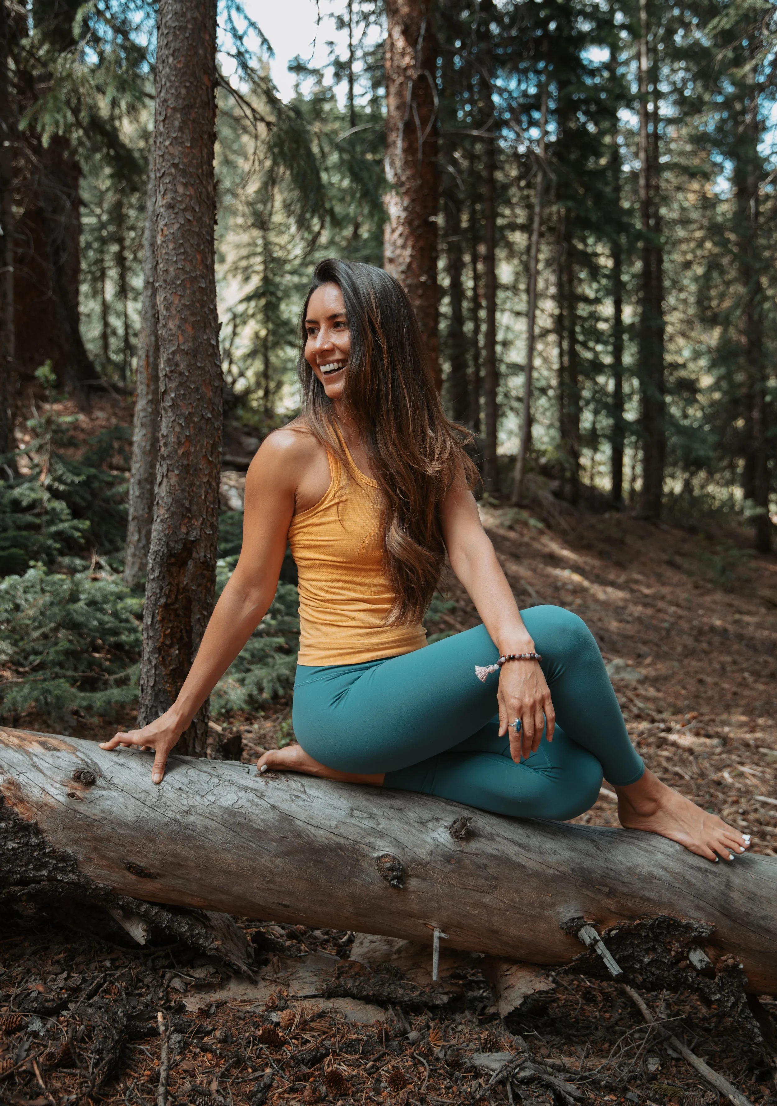 A smiling woman with long dark hair, wearing a yellow tank top and teal yoga pants, sitting on a fallen tree trunk in a forest surrounded by trees and greenery.