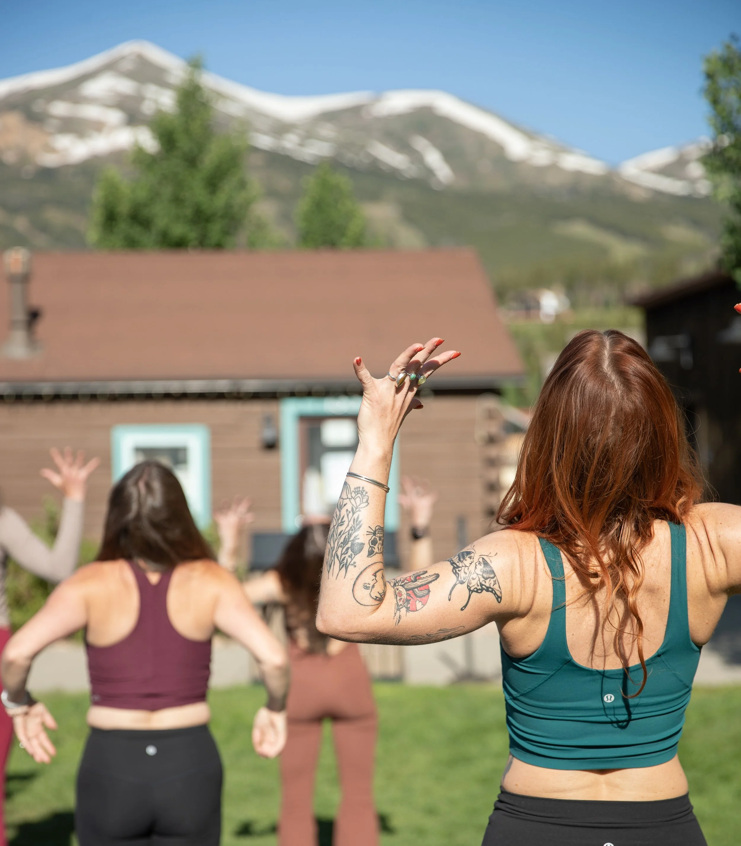 Group of women practicing yoga outdoors with mountains in background, focusing on woman with long red hair in teal sports bra raising hand, other women in background in different colored sports bras raising hands.