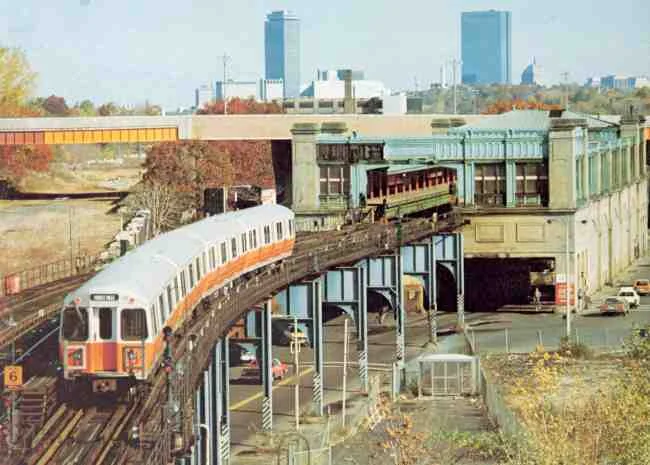 The Orange Line MBTA train reaches the end of the line at Forest Hills in this 1980s photo. The Arborway overpass can be seen along with the distant views of the Prudential and John Hancock buildings.