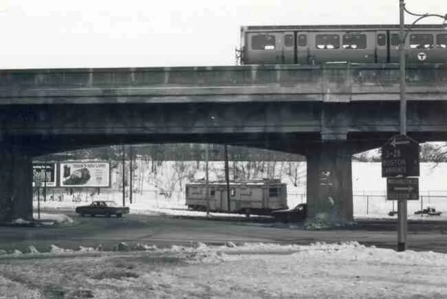 A service trolley heads toward South Street. The overhead train leaves Forest Hills Station on its way to Boston in this 1968 photo taken at the Washington Street rotary. The top street sign points toward Route 3-28 and reads Boston 4 and Lawrence 33. The bottom sign reads Washington Street and Egleston Square.