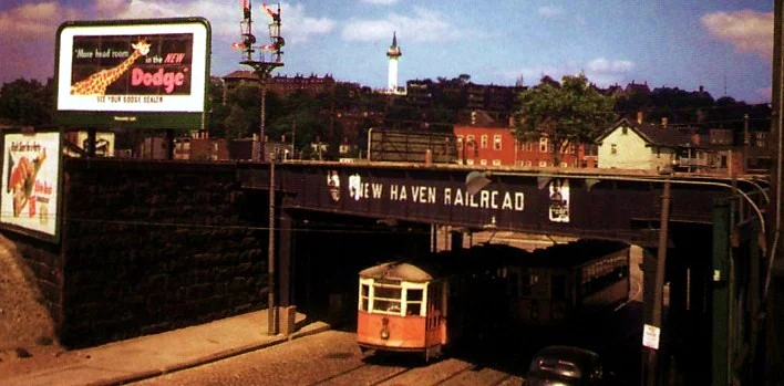 This 1947 photograph shows a trolley passing in each direction on Centre St. under the railroad bridge at Jackson Square. The standpipe on Fort Hill can be seen in the background.