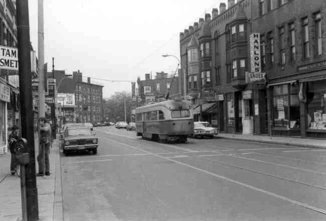 A lone trolley passes the Burroughs Building on its way down Centre Street towards the Arborway in this 1960 photo. Established businesses at the time were Rogers Drug Store, Frank’s DoubleWare Clothing, Barry’s Deli, Blanchard Liquors, and Galway House Restaurantand Hanlons Shoes. 