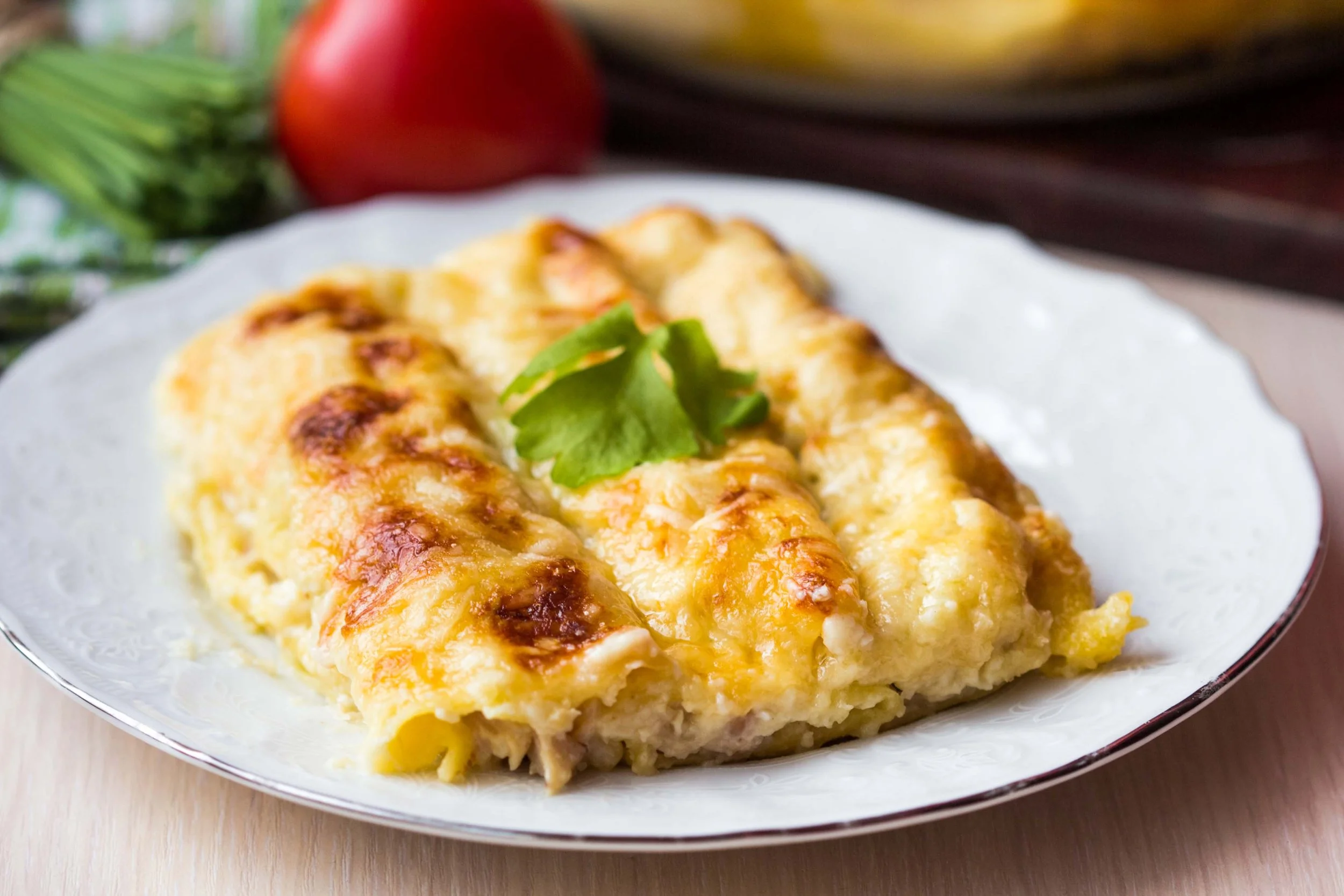 Baked cheesy casserole garnished with a cilantro leaf on a white plate, with a tomato, zucchini, and bananas in the background.